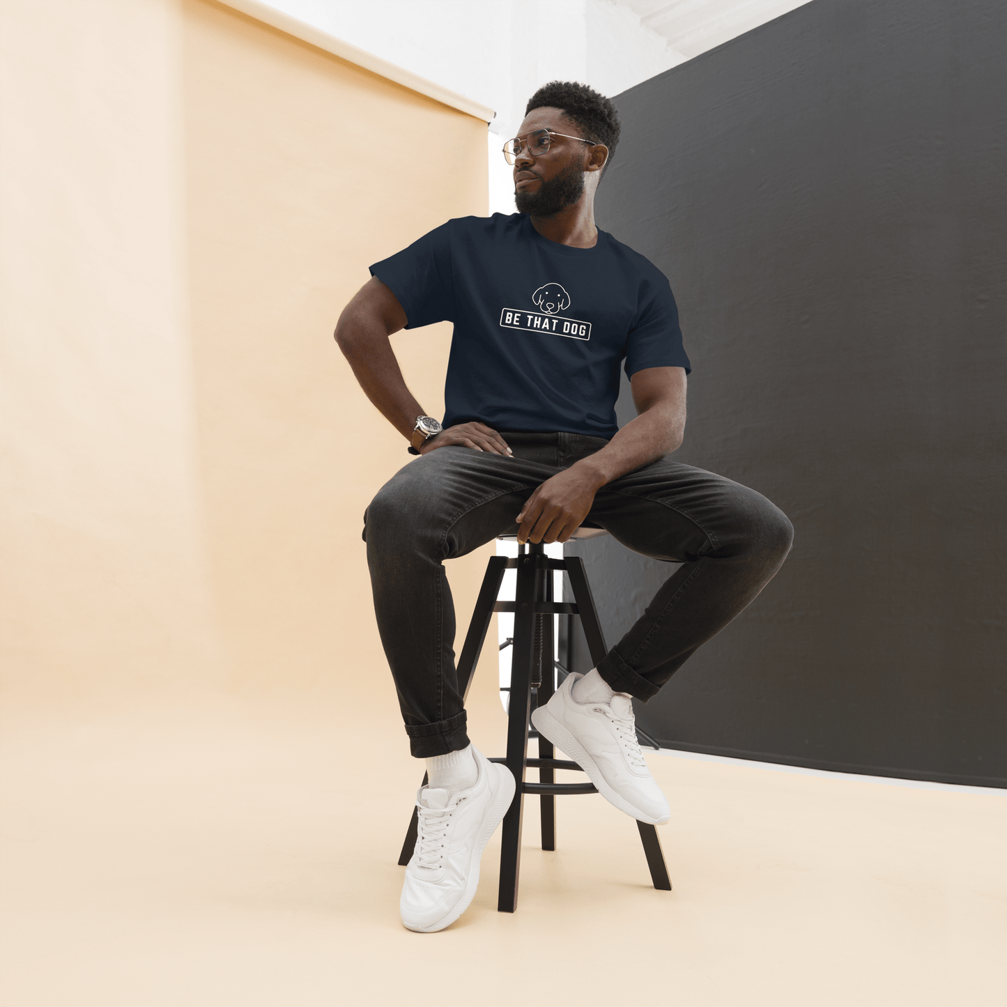 A man wearing glasses and a dark "Be THAT Dog Minimalist Pup Face Tee" by THiNK LiKE A DOG®, made from comfy cotton, sits on a black stool against a minimalist studio backdrop.