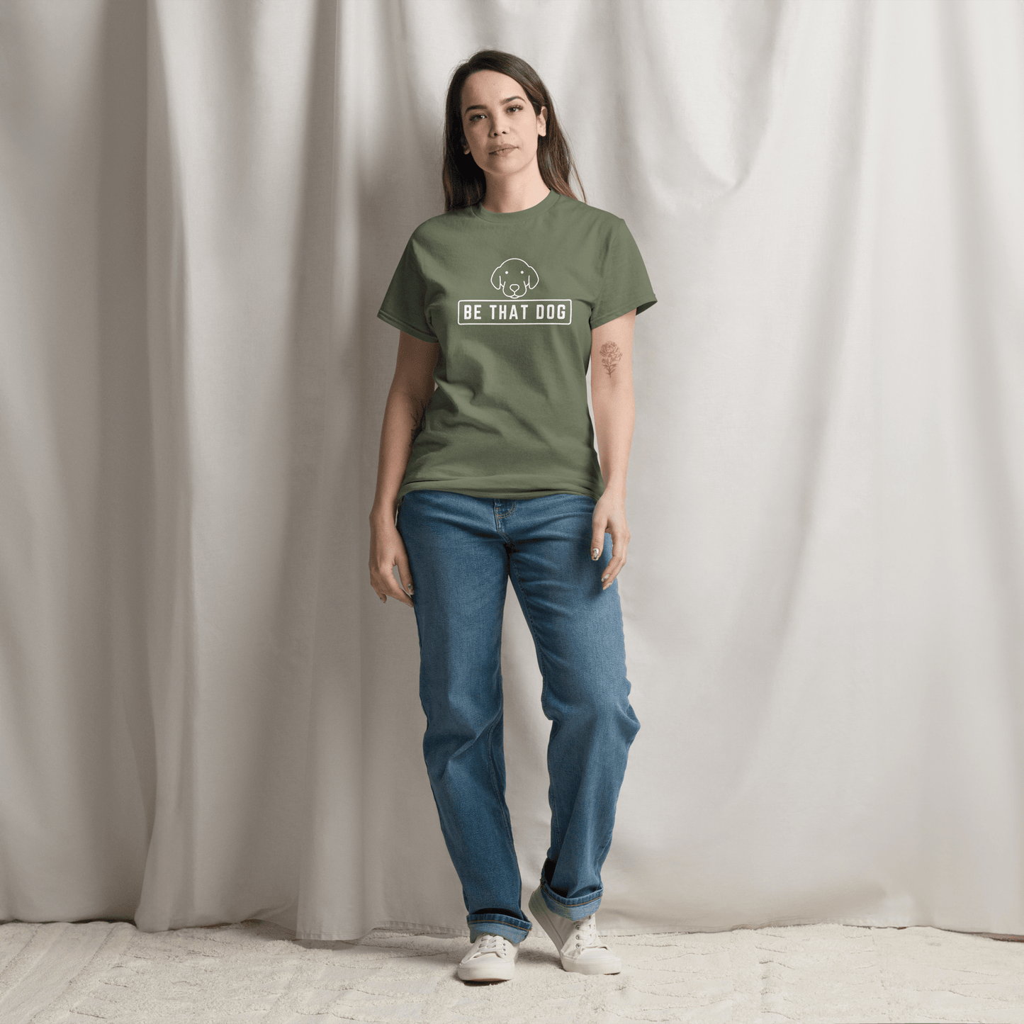 A person in a green "Be THAT Dog Minimalist Pup Face Tee" by THiNK LiKE A DOG®, made from comfortable cotton with a simple dog design, stands in front of a light curtain.