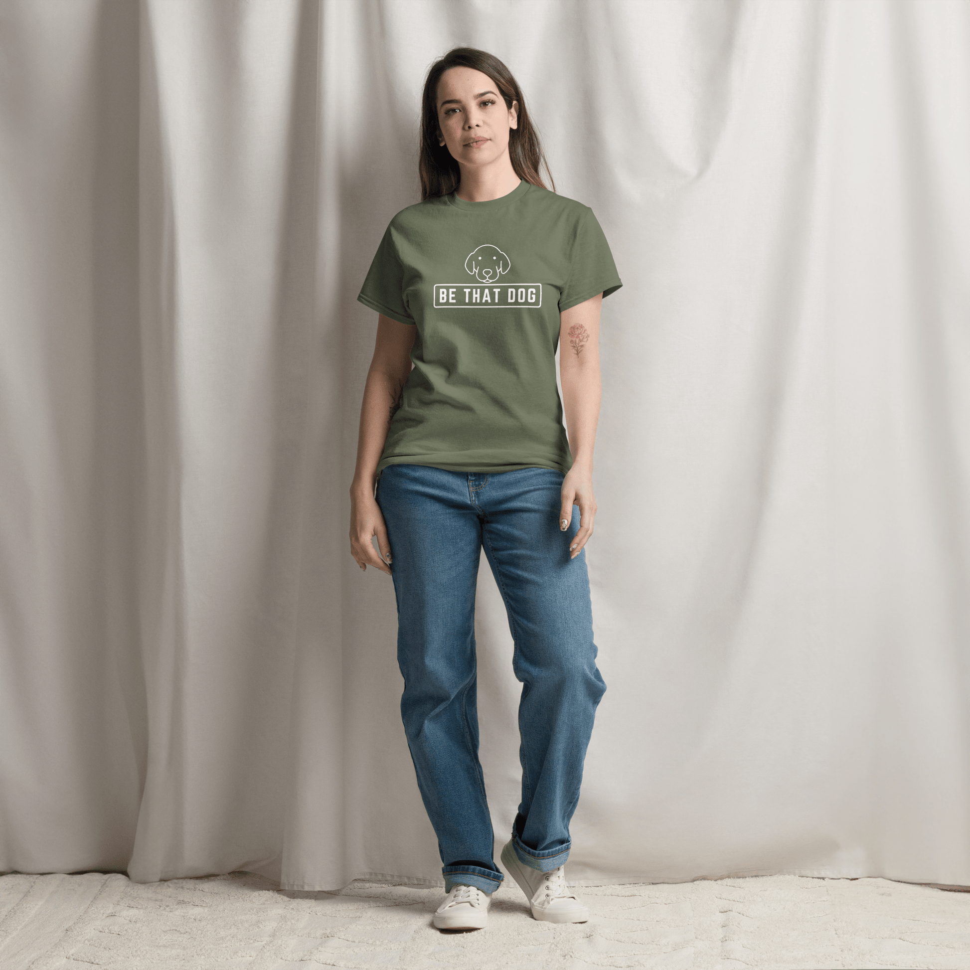 A person in a green "Be THAT Dog Minimalist Pup Face Tee" by THiNK LiKE A DOG®, made from comfortable cotton with a simple dog design, stands in front of a light curtain.