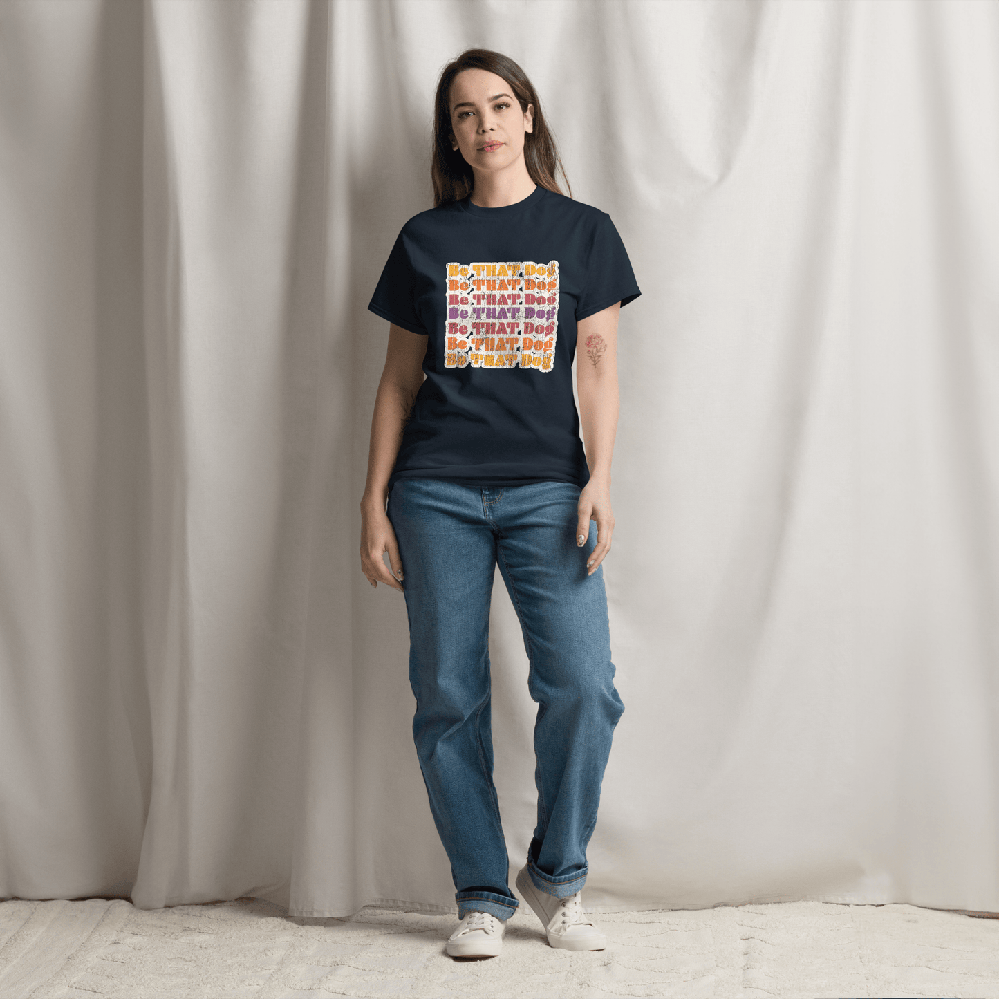 A person stands on a carpet, wearing blue jeans and the THiNK LiKE A DOG® Be That Dog Retro Unisex Tee with colorful text, in front of a draped white curtain.