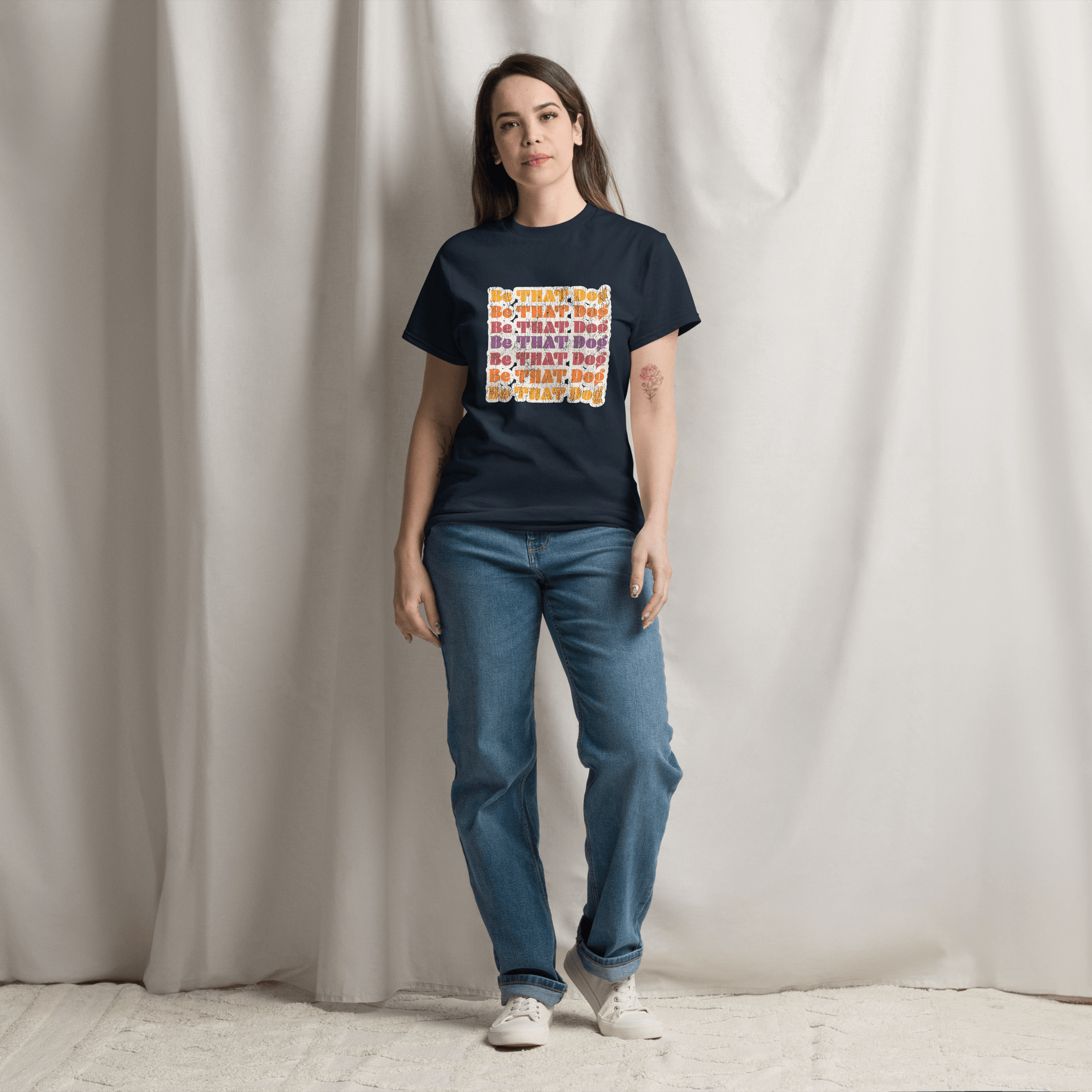 A person stands on a carpet, wearing blue jeans and the THiNK LiKE A DOG® Be That Dog Retro Unisex Tee with colorful text, in front of a draped white curtain.