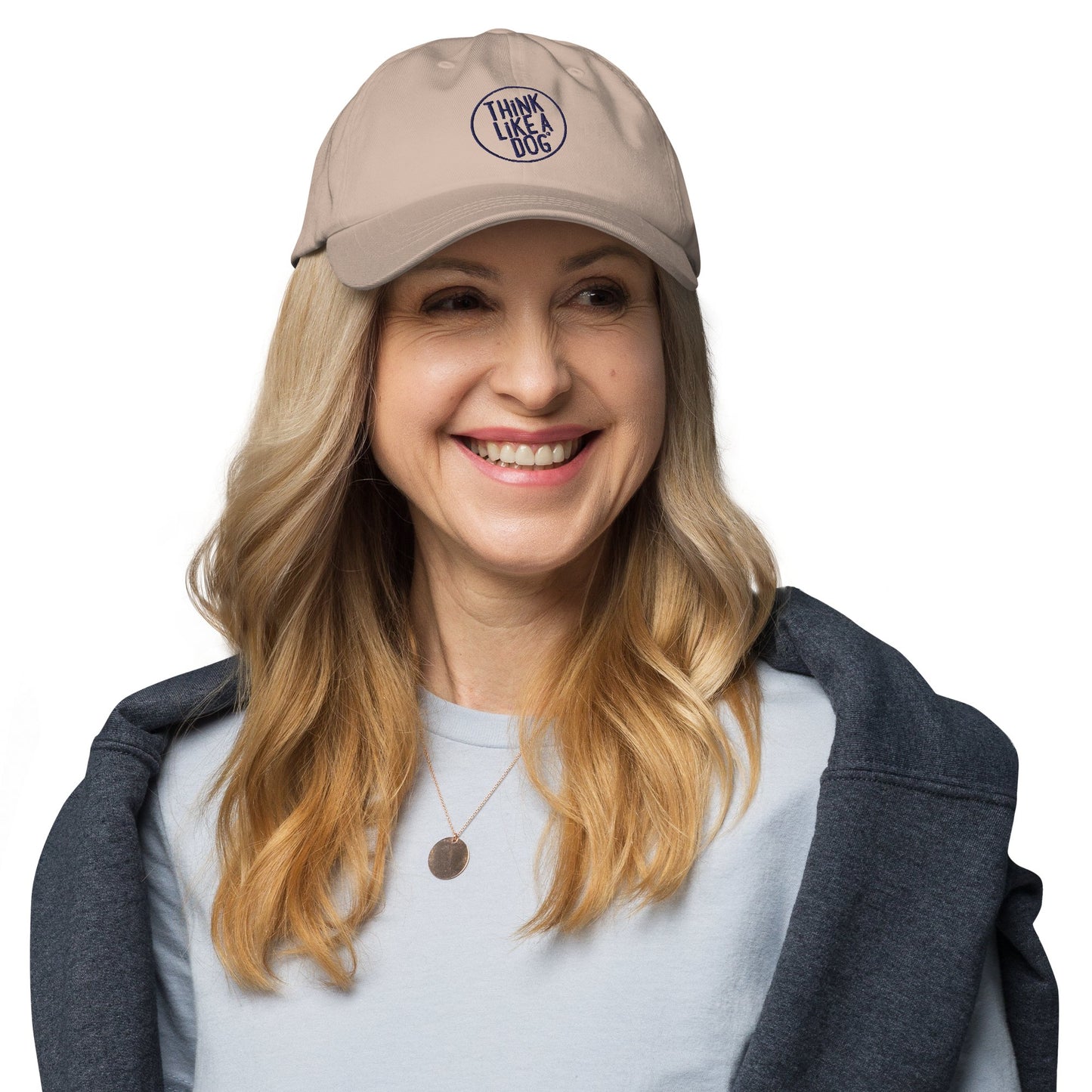 A woman with long blonde hair smiles while wearing the Classic Navy Dad Hat featuring the "Think Like a Dog" circle logo by THiNK LiKE A DOG®. As a dog lover, she is dressed in a light blue shirt and has a dark jacket draped over her shoulders.