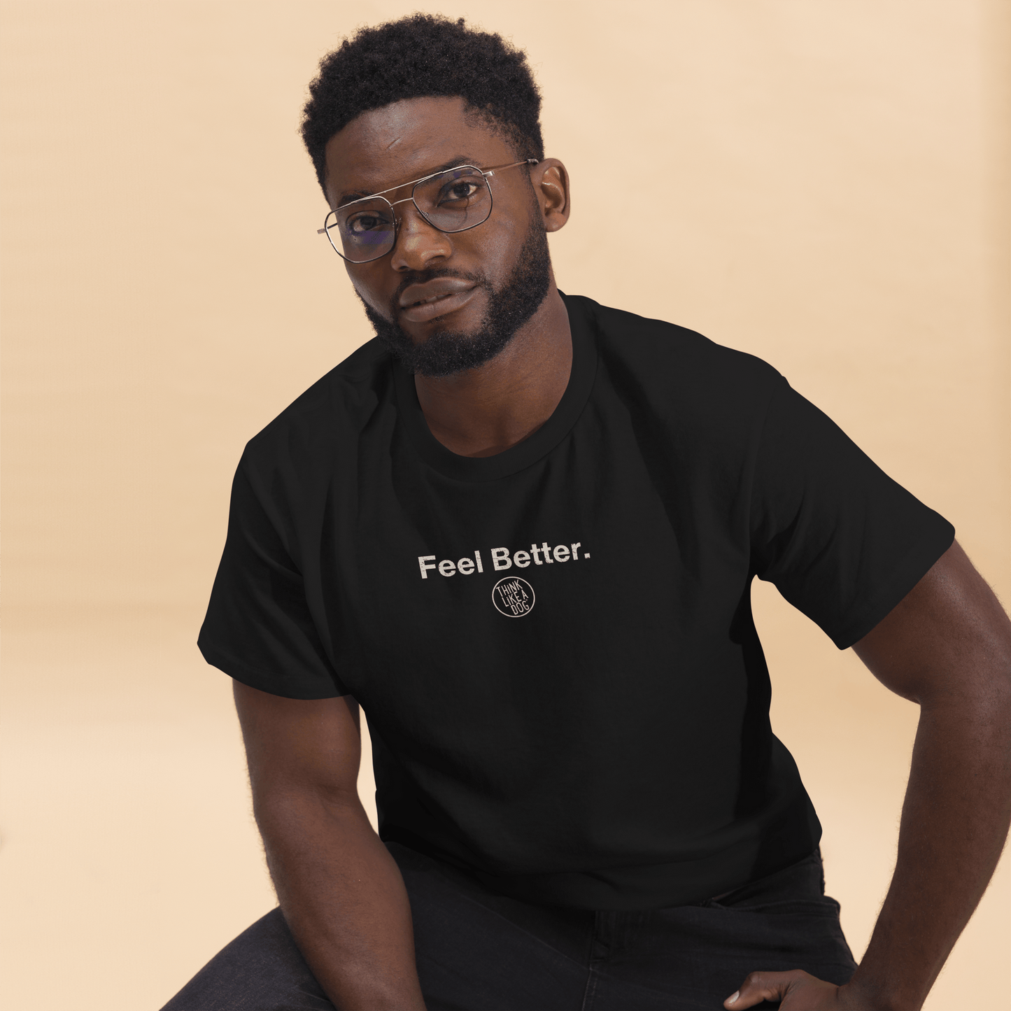A man in a black "Feel Better Dog Lover T-Shirt with Logo" by THiNK LiKE A DOG® sits against a beige background, exuding the calm confidence of someone who fully embraces the mantra.