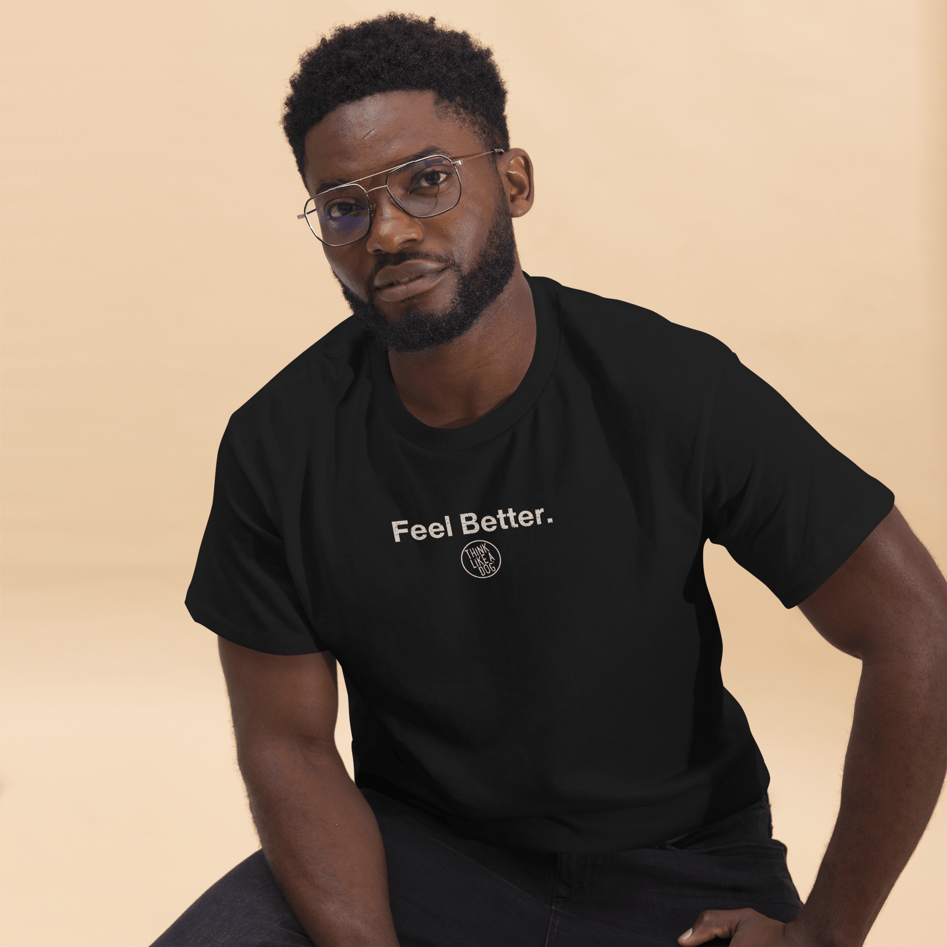 A man in a black "Feel Better Dog Lover T-Shirt with Logo" by THiNK LiKE A DOG® sits against a beige background, exuding the calm confidence of someone who fully embraces the mantra.