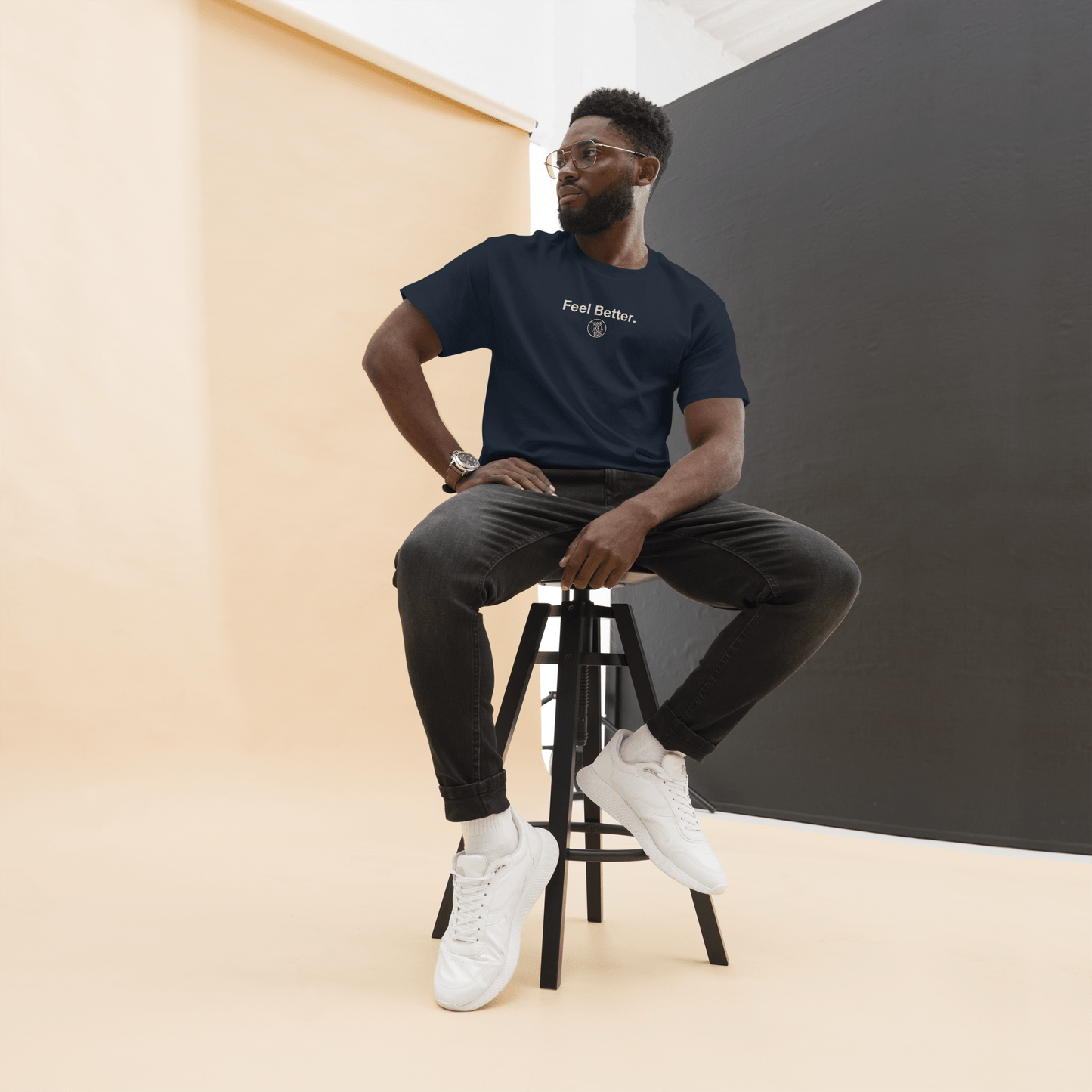 A man with glasses sits on a stool in a studio with beige and black walls, wearing dark jeans, white sneakers, and the THiNK LiKE A DOG® Feel Better Dog Lover T-Shirt with Logo—a comforting gift for pet owners—while looking to the side.