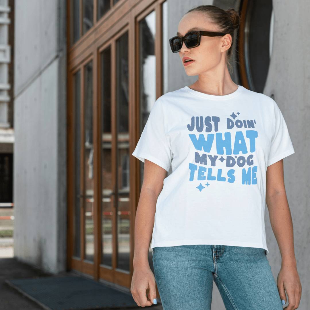 A woman in sunglasses wears the THiNK LiKE A DOG® "Just Doin' What My Dog Tells Me — Bold Tee," a white unisex shirt with…