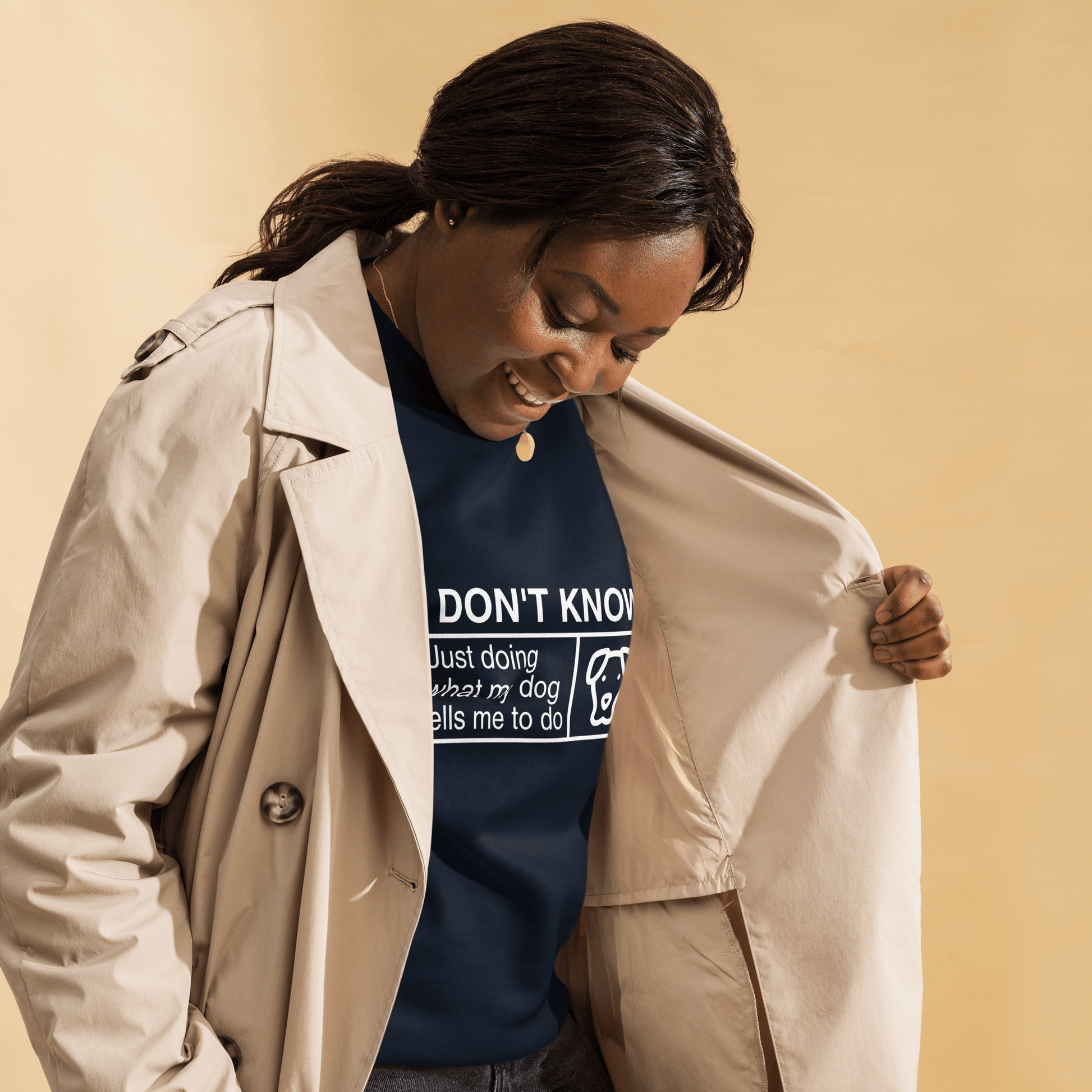 With a beige coat open, someone reveals a navy "I Don’t Know, Just Doing What My Dog Tells Me" sweatshirt by THiNK LiKE A DOG®, smiling against a beige background.