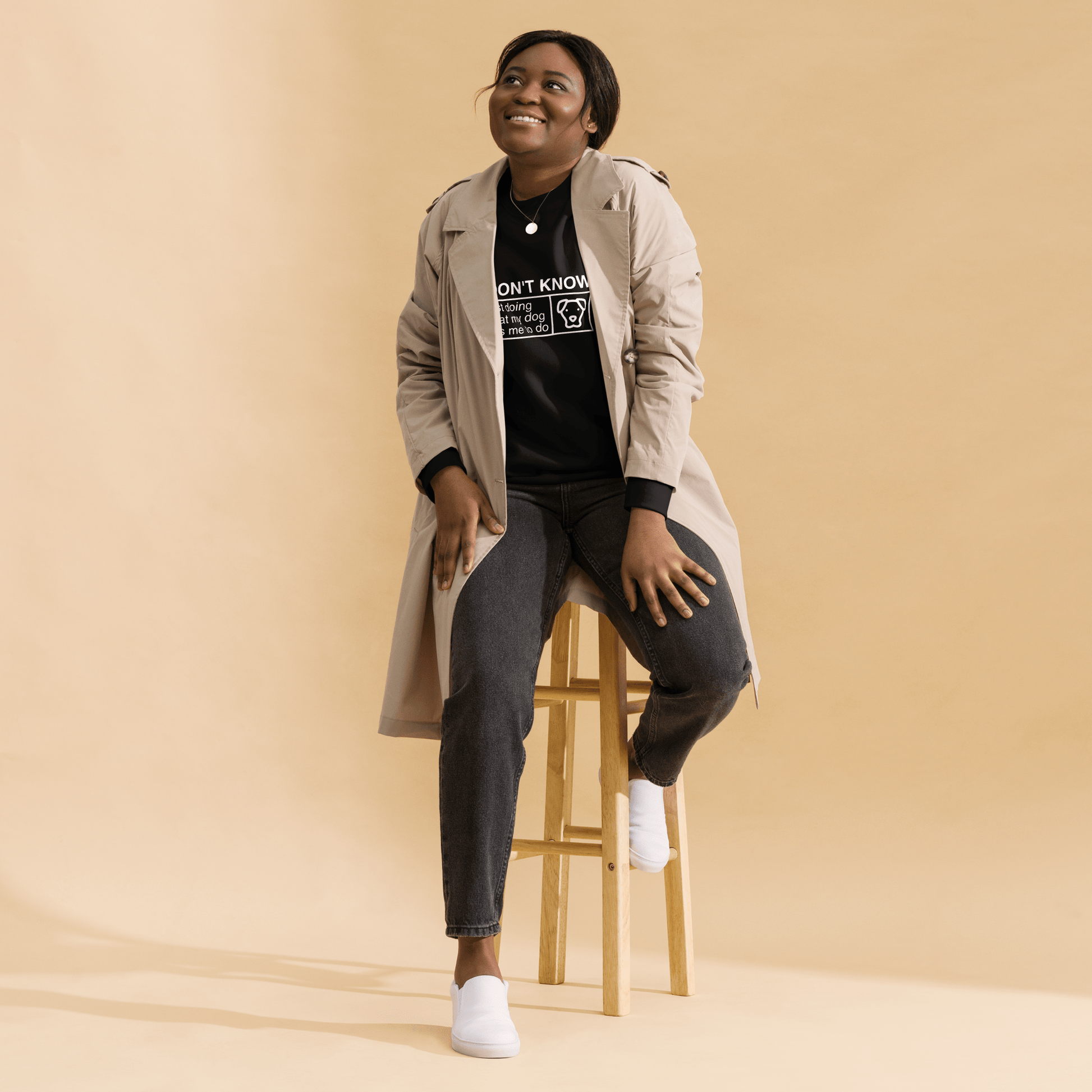 A person smiles on a wooden stool, wearing the THiNK LiKE A DOG® "I Don’t Know, Just Doing What My Dog Tells Me" sweatshirt, dark jeans, and white shoes against a beige background.