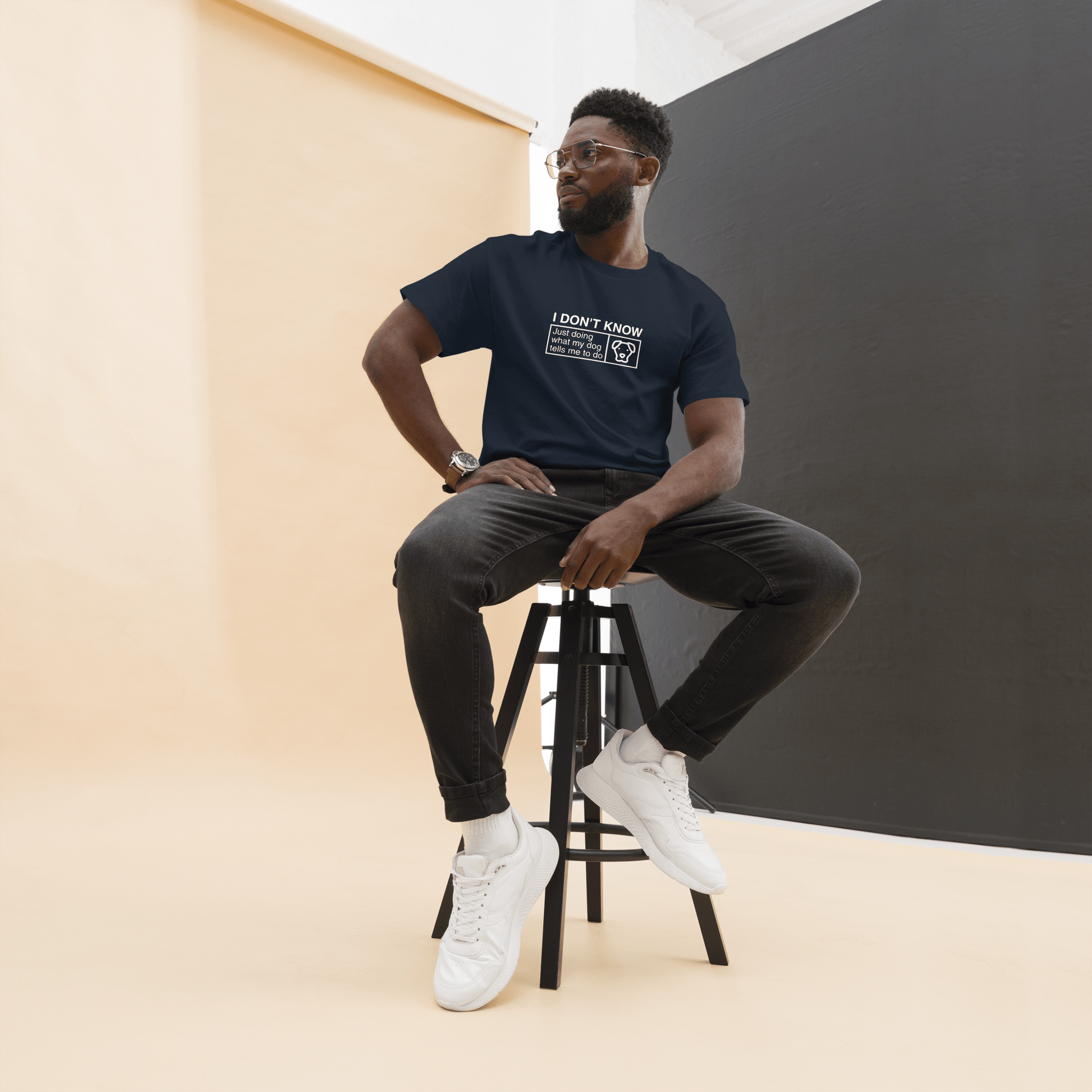 A man sits on a tall stool in a studio, wearing the THiNK LiKE A DOG® Funny Dog Shirt – Dog Icon “Just Doing What My Dog Tells Me,” paired with dark jeans, white sneakers, and glasses against a neutral two-tone background.
