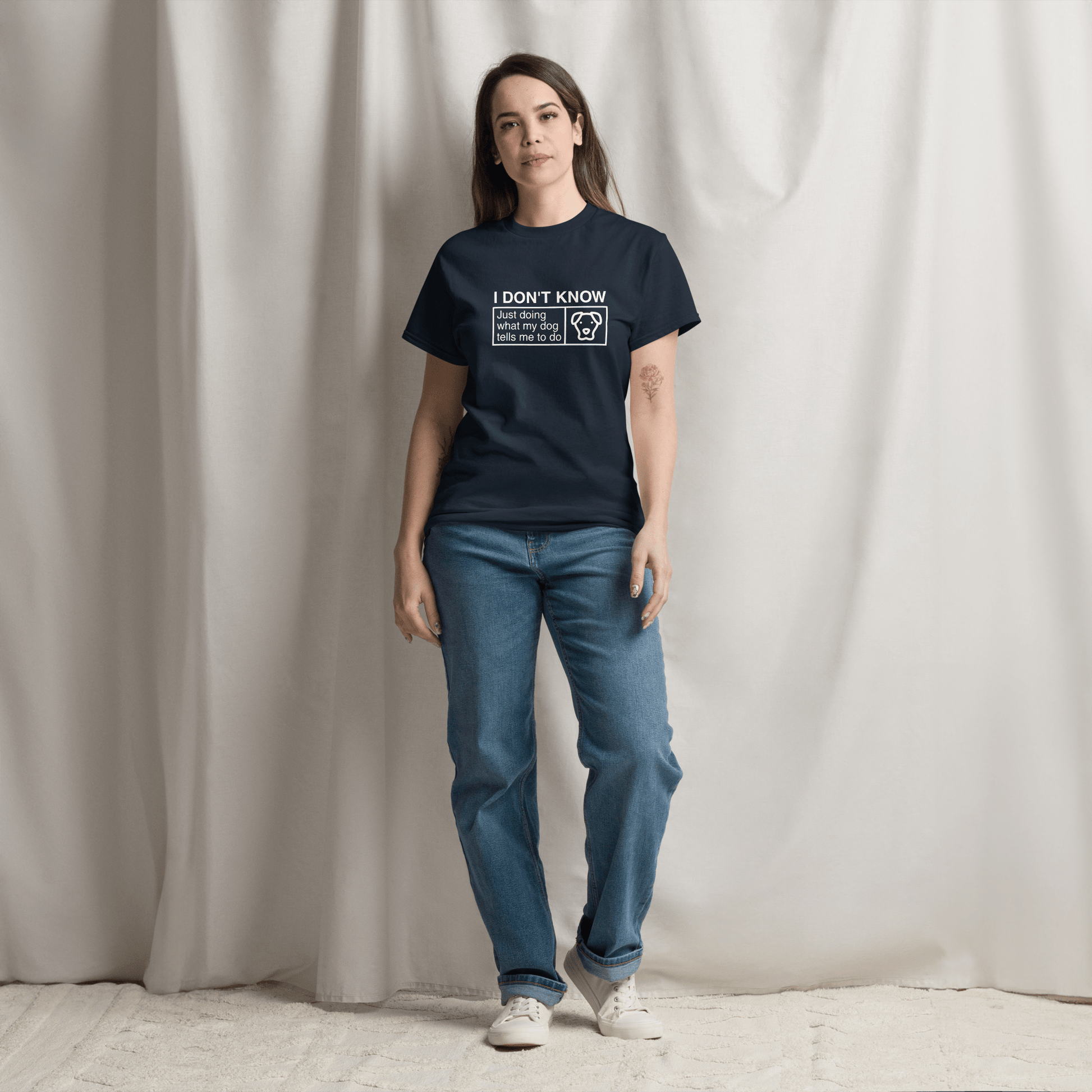 A woman stands before beige curtains wearing a navy Funny Dog Shirt by THiNK LiKE A DOG® featuring the text “Just Doing What My Dog Tells Me” with a dog icon and blue jeans—ideal as a gift for dog lovers.