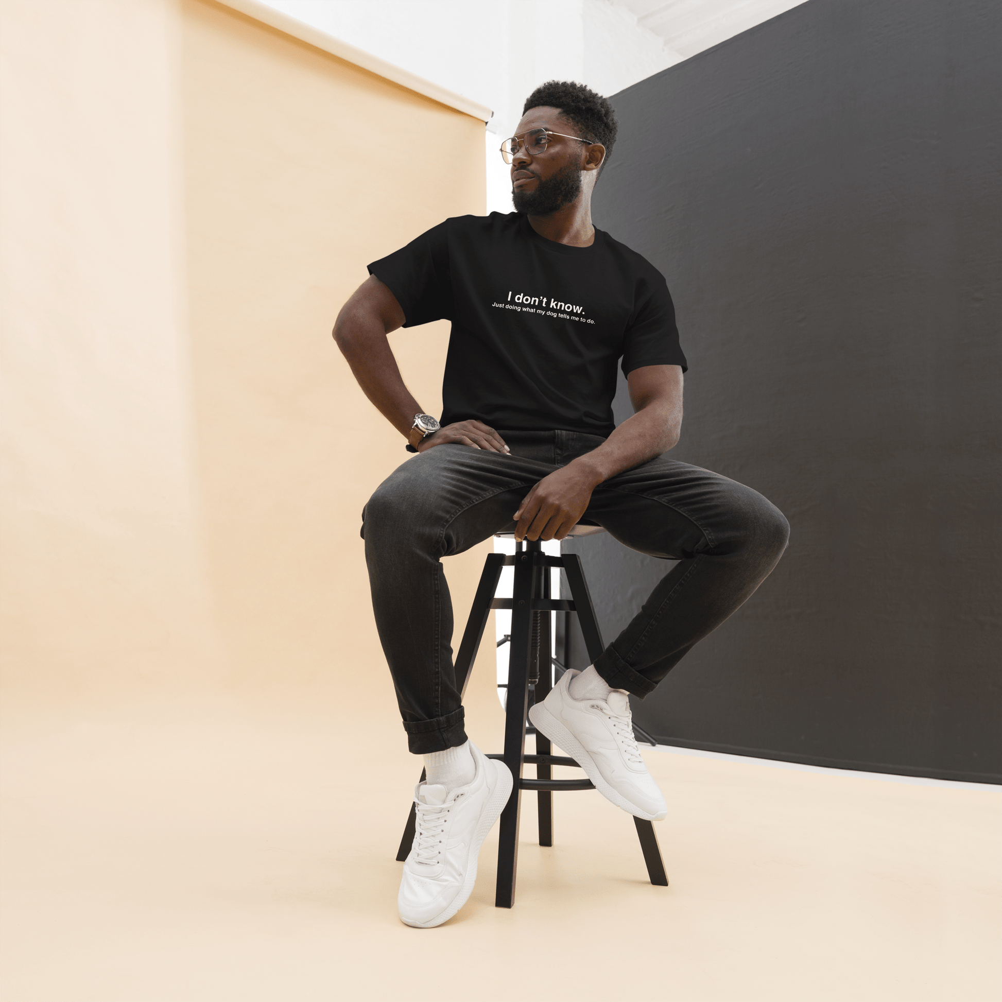 A man with glasses sits on a stool in a studio setting with beige and dark gray backgrounds, sporting black jeans, white sneakers, and THiNK LiKE A DOG®'s Funny Dog Shirt - "Just Doing What My Dog Tells Me," adding a humorous twist to dog apparel.