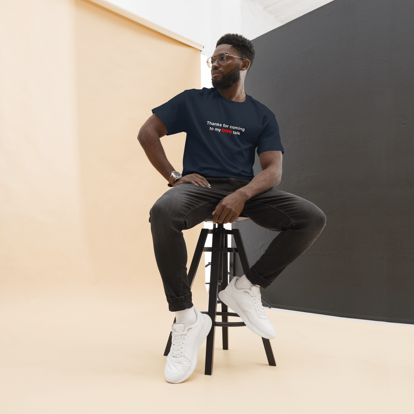 A man wearing glasses and a THiNK LiKE A DOG® "Thanks for Coming to My Dog Talk" funny dog shirt sits on a tall stool against a beige and black studio background—a perfect gift for dog lovers.