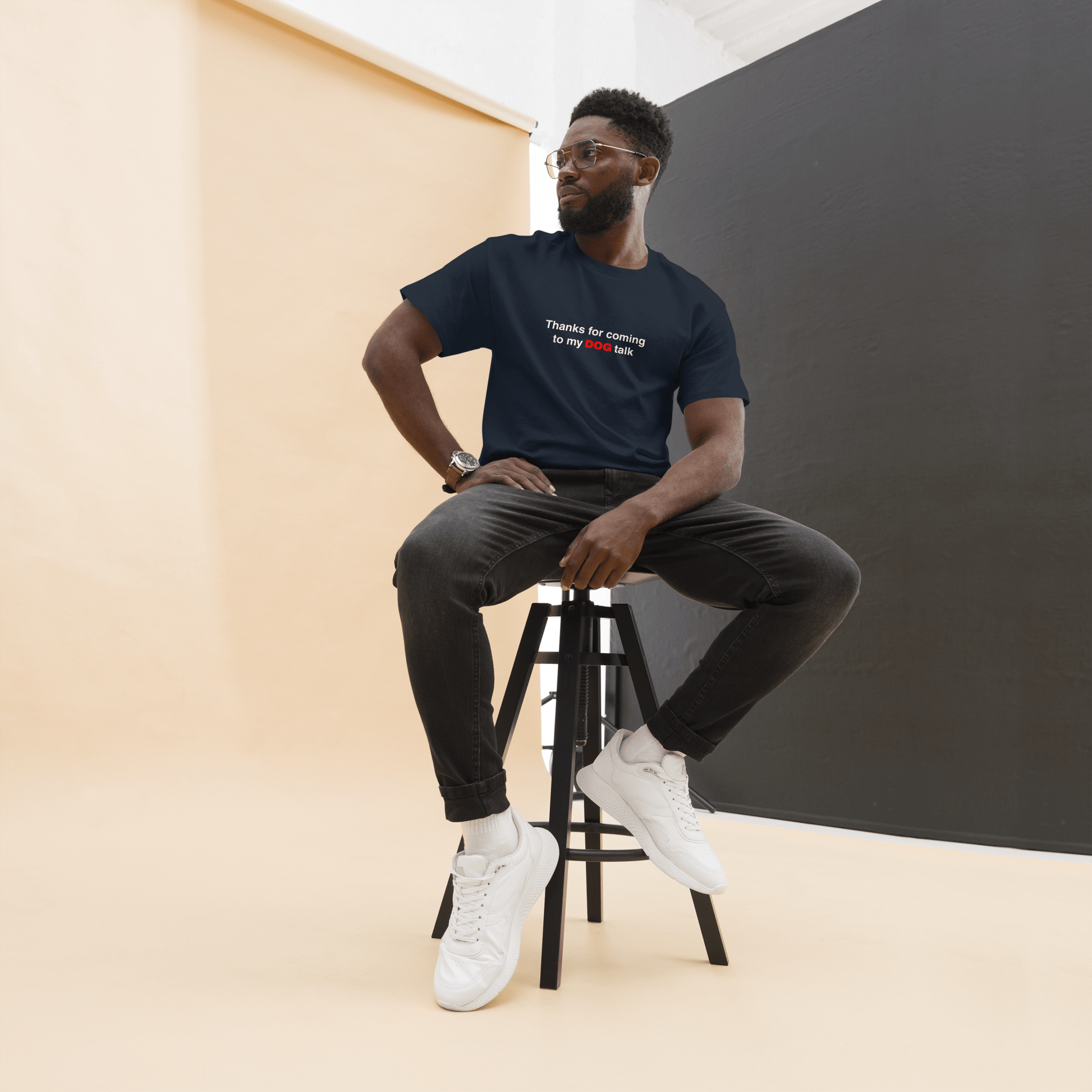 A man wearing glasses and a THiNK LiKE A DOG® "Thanks for Coming to My Dog Talk" funny dog shirt sits on a tall stool against a beige and black studio background—a perfect gift for dog lovers.