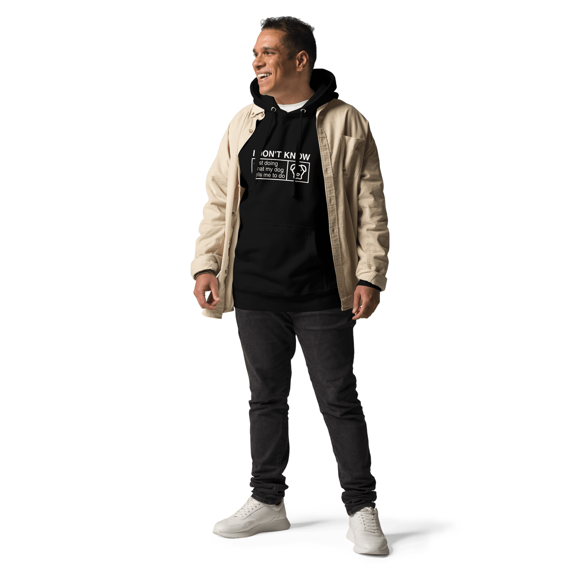 A person smiles, wearing a beige jacket, the THiNK LiKE A DOG® "I Don’t Know, Just Doing What My Dog Tells Me" hoodie, dark jeans, and white sneakers against a black background.