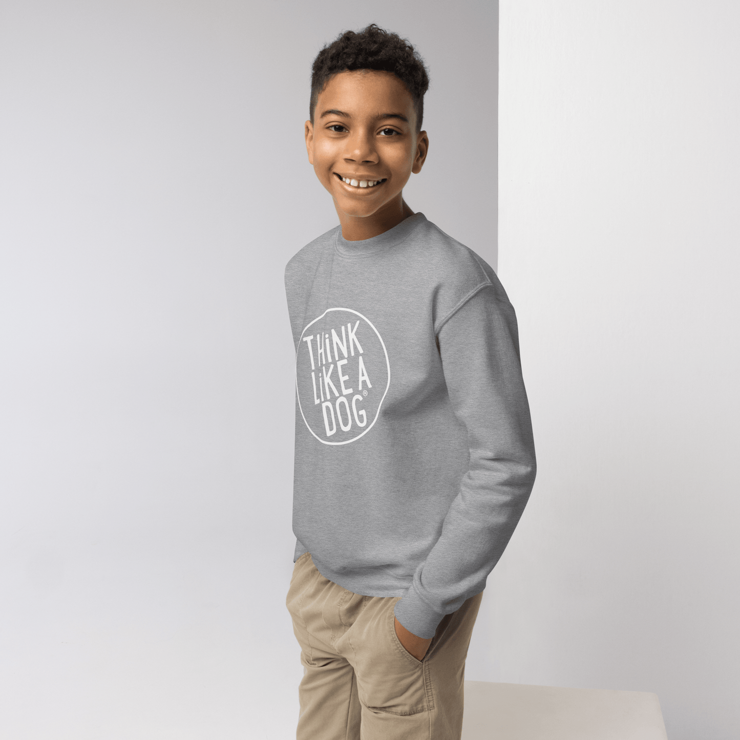 A young person beams in a cozy crewneck, featuring the white "THiNK LiKE A DOG®" logo. Perfect for kids who love dogs, this Kids' Cozy Crewneck Sweatshirt by THiNK LiKE A DOG® stands out against the light background.