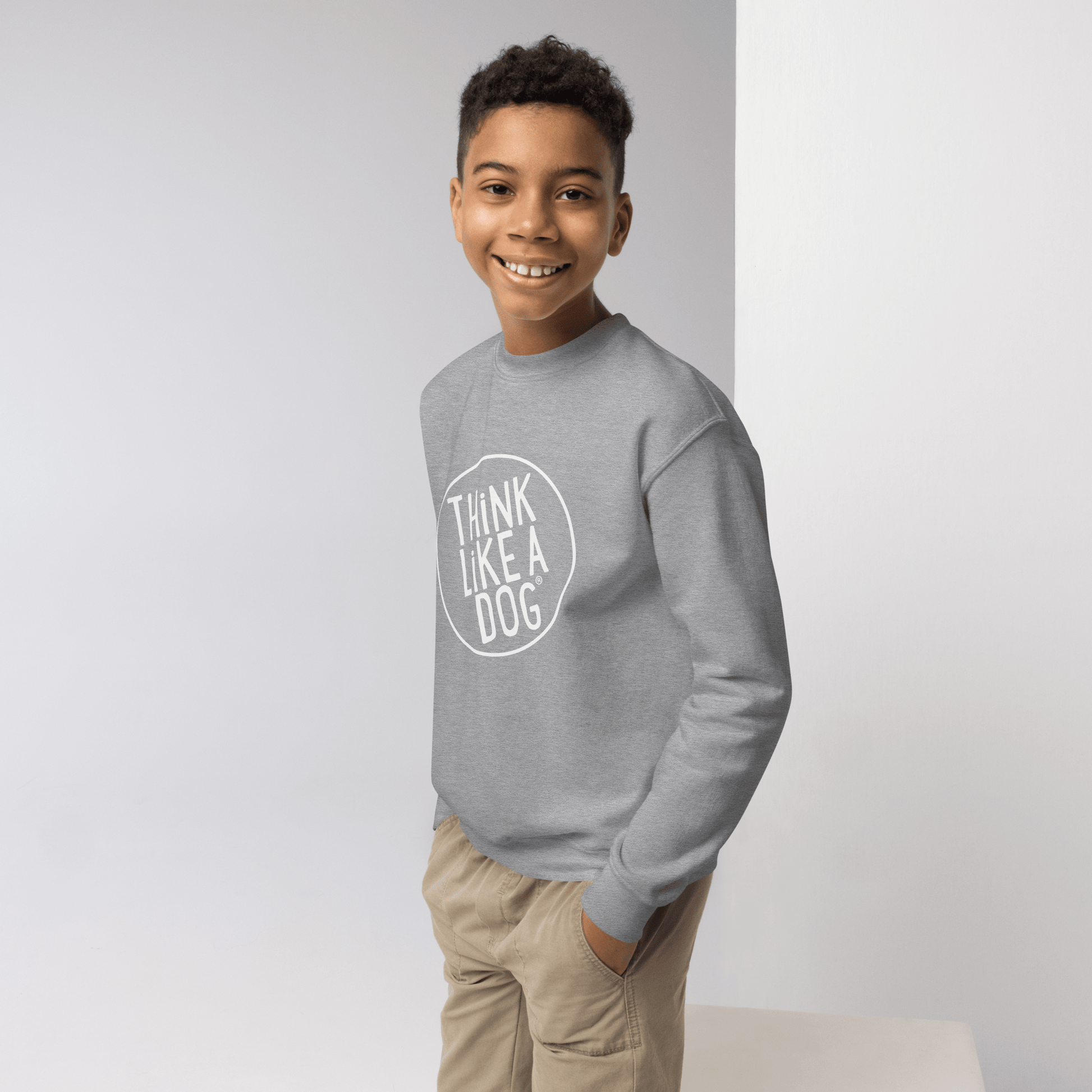 A young person beams in a cozy crewneck, featuring the white "THiNK LiKE A DOG®" logo. Perfect for kids who love dogs, this Kids' Cozy Crewneck Sweatshirt by THiNK LiKE A DOG® stands out against the light background.