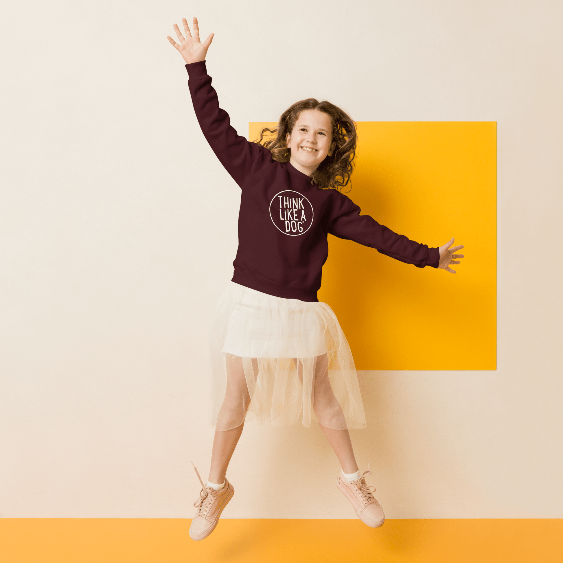 A girl joyfully jumps against a beige and orange backdrop, wearing a maroon Kids' Cozy Crewneck Sweatshirt featuring the white THiNK LiKE A DOG® logo. Her outfit includes a white skirt and blush sneakers, earning the Dog-Lover Approved tag.