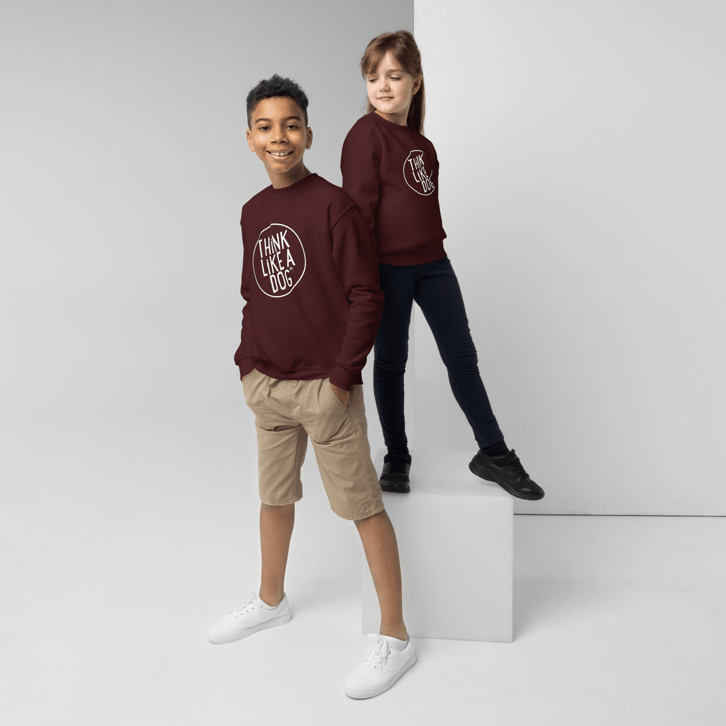 Two kids create a delightful scene in Kids' Cozy Crewneck Sweatshirts by THiNK LiKE A DOG® with the white logo. One stands confidently on the floor wearing beige shorts and white shoes, while the other perches on a white cube in dark pants and black shoes.