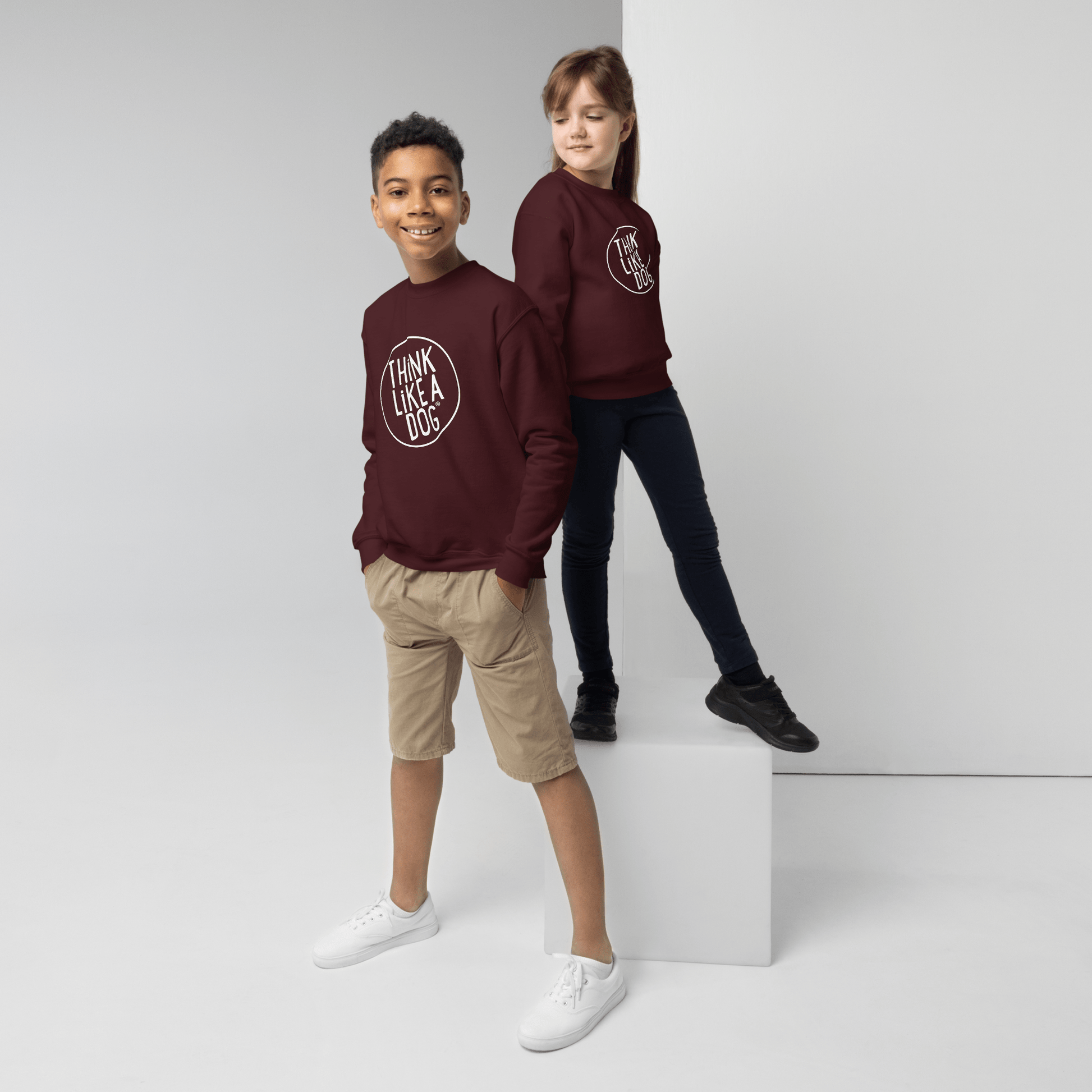 Two kids create a delightful scene in Kids' Cozy Crewneck Sweatshirts by THiNK LiKE A DOG® with the white logo. One stands confidently on the floor wearing beige shorts and white shoes, while the other perches on a white cube in dark pants and black shoes.