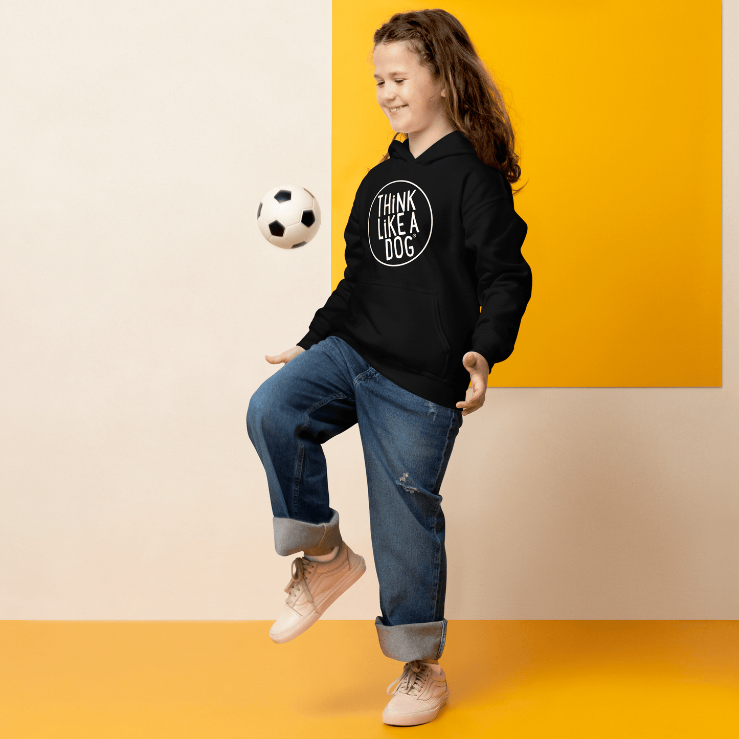 A dog-lover style shines as a person in a black Kids Heavy Blend Hoodie featuring the THiNK LiKE A DOG® white logo, paired with blue jeans, skillfully juggles a soccer ball indoors against a yellow and beige wall.