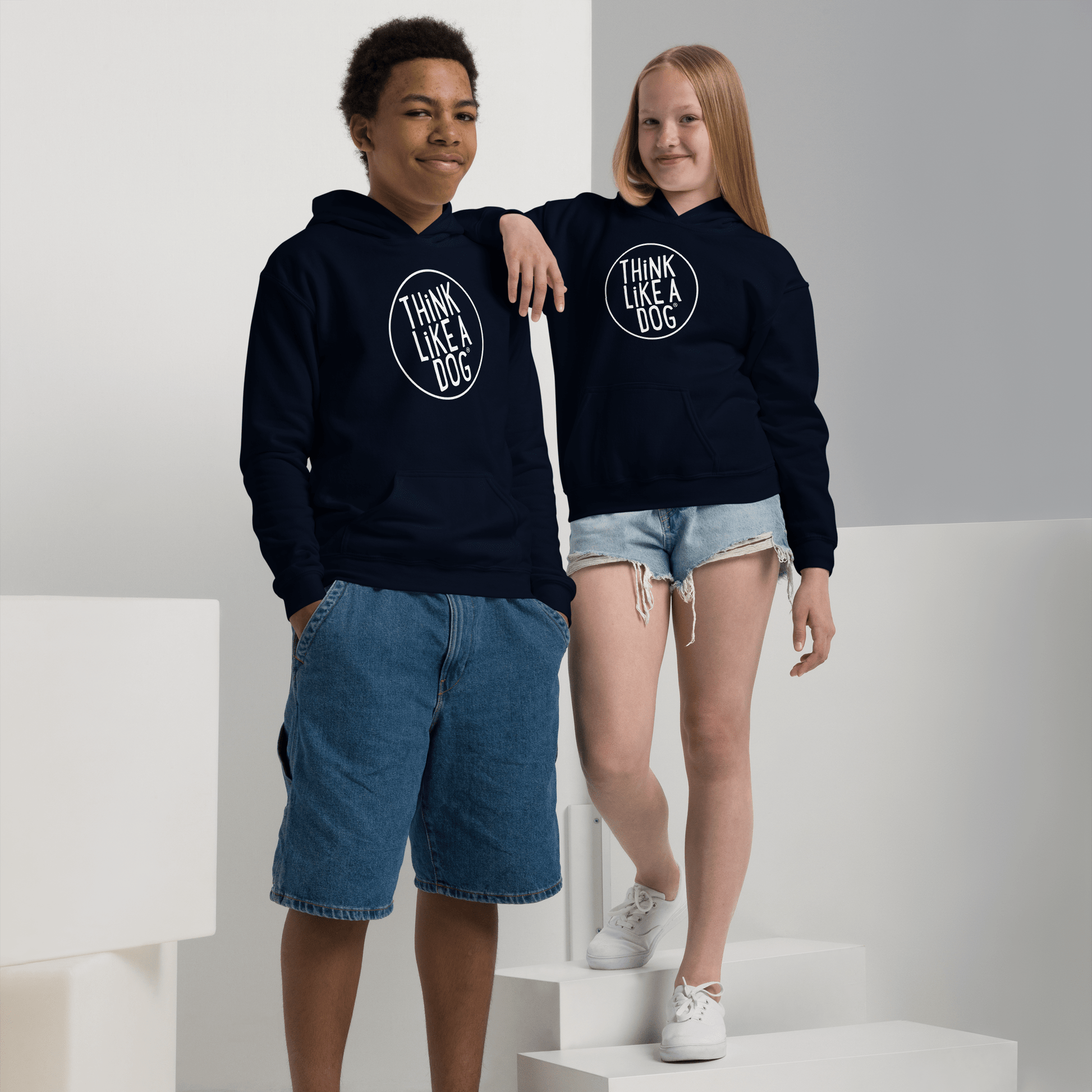 Two people wear navy Kids Heavy Blend Hoodies featuring the THiNK LiKE A DOG® White Logo. One pairs it with denim shorts and hands in pockets, while the other stands on a white block, fully embracing their Dog-Lover Style—perfect for fans of cozy and stylish hoodies.