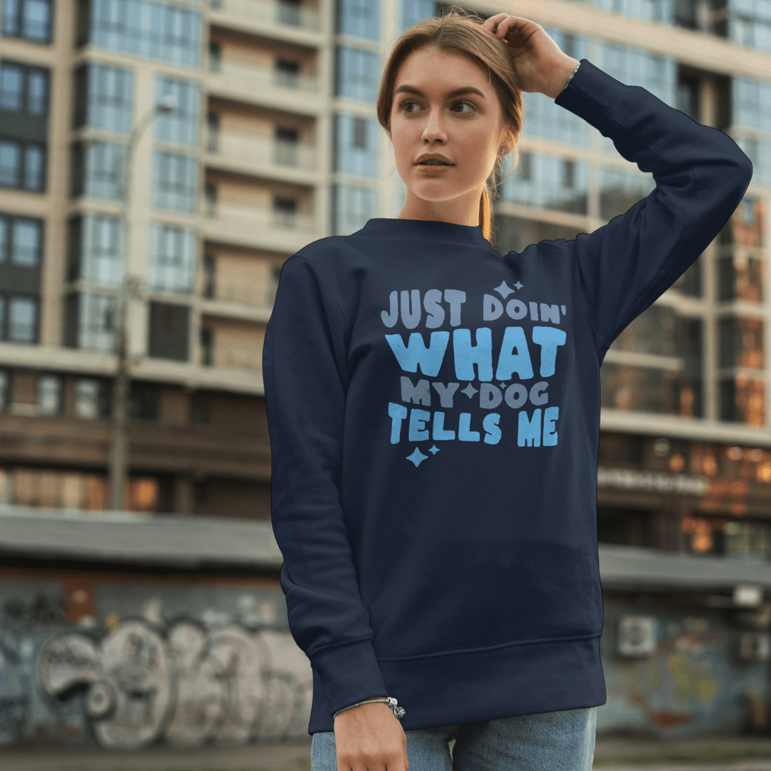 A woman stands outdoors in front of a building, wearing the THiNK LiKE A DOG® Think Like a Dog Sweatshirt — Everyday Comfort, featuring "Just doin' what my dog tells me" in blue and white—a perfect pick for dog lovers.