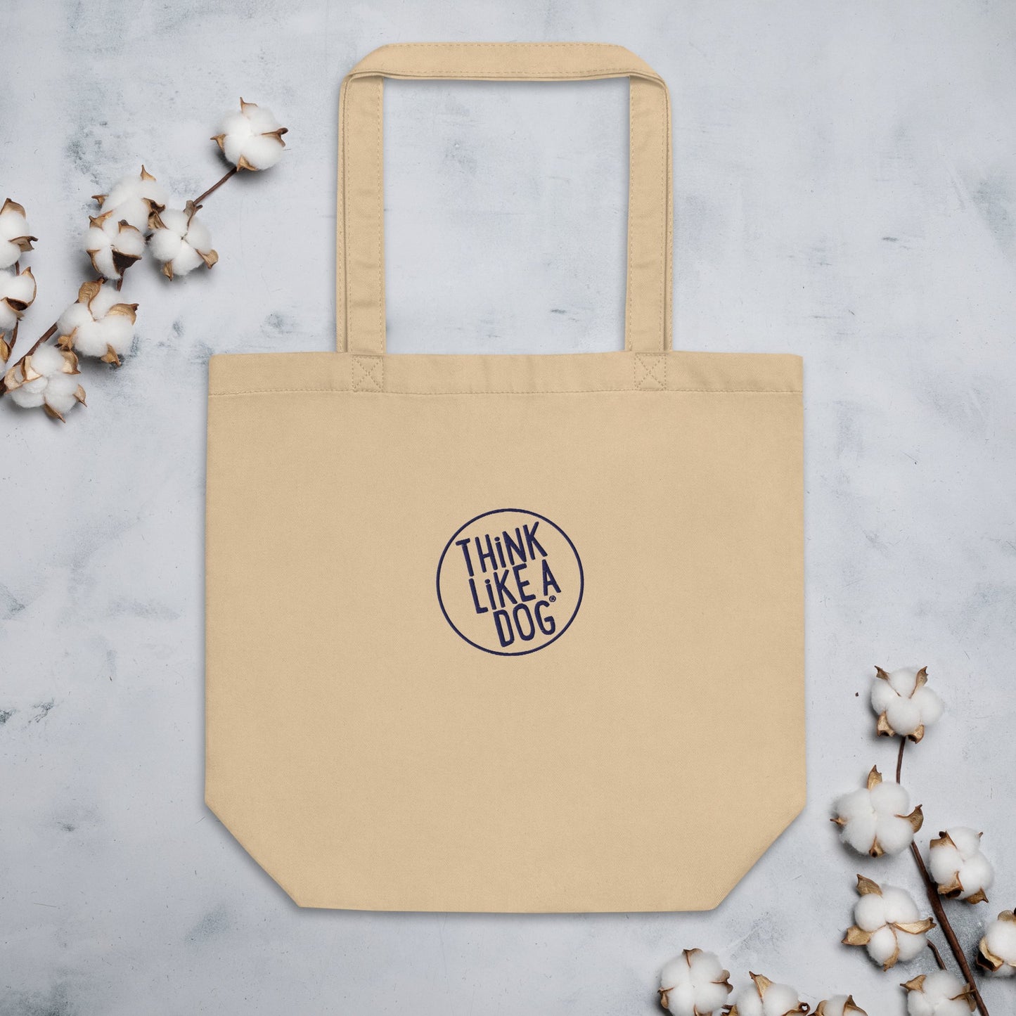 A stylish and sustainable THiNK LiKE A DOG® Eco-Friendly Beige Tote Bag with the "Think Like a Dog" embroidered logo inside a circular border is placed on a marble surface, flanked by cotton branches on both sides. Made from durable materials, this tote is perfect for the eco-conscious shopper.