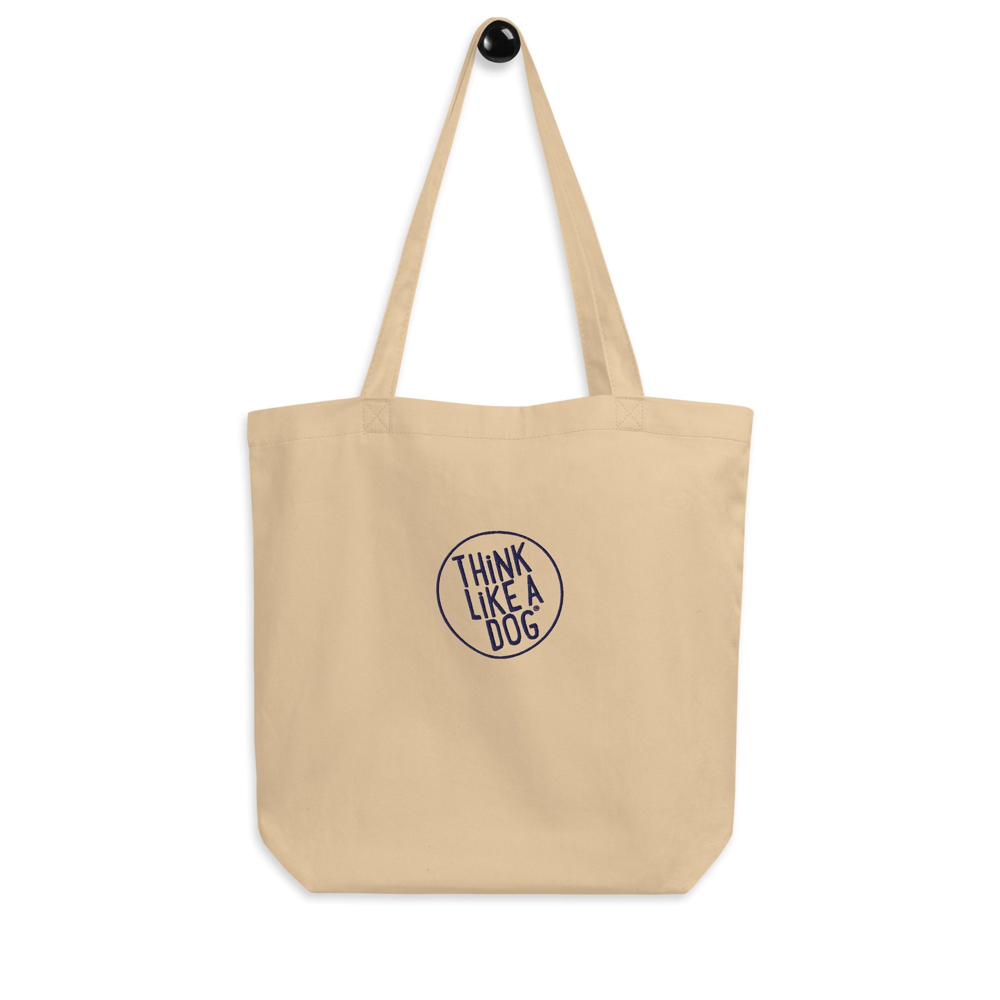 A THiNK LiKE A DOG® Eco-Friendly Beige Tote Bag, featuring a durable design and an embroidered circular logo with the text "THINK LIKE A DOG," hangs on a wall hook.