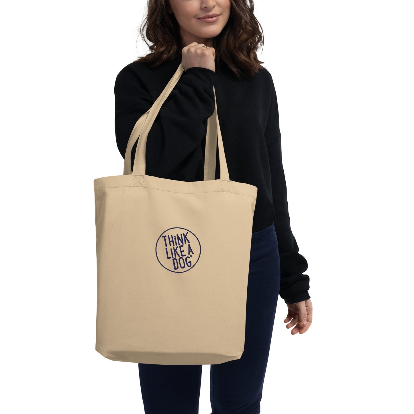 A person holding a THiNK LiKE A DOG® Eco-Friendly Beige Tote Bag with the "Think Like a Dog" embroidered logo, wearing a black top and navy pants.