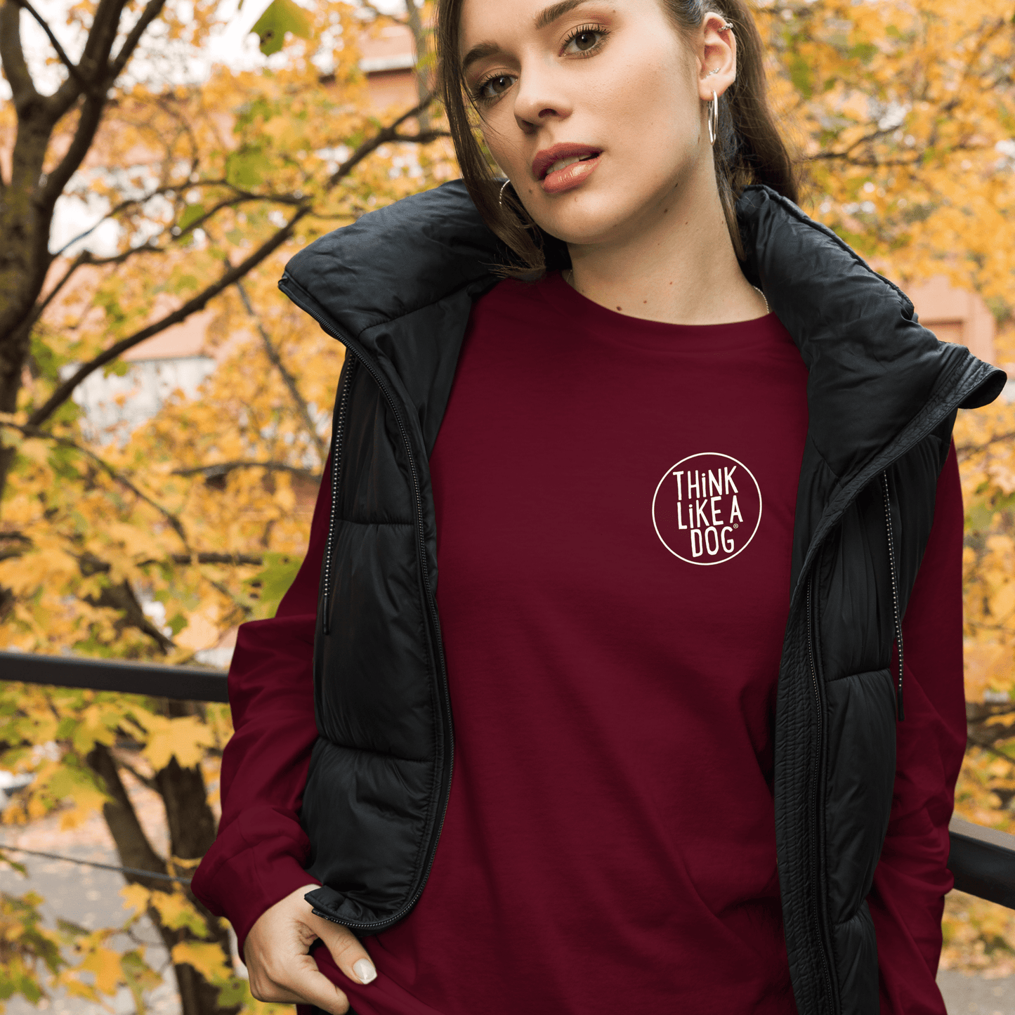 A person in a maroon "THiNK LiKE A DOG Long Sleeve Tee | Minimalist Dog Lover Shirt" by THiNK LiKE A DOG® is outside wearing a black vest, with autumn leaves in the background.