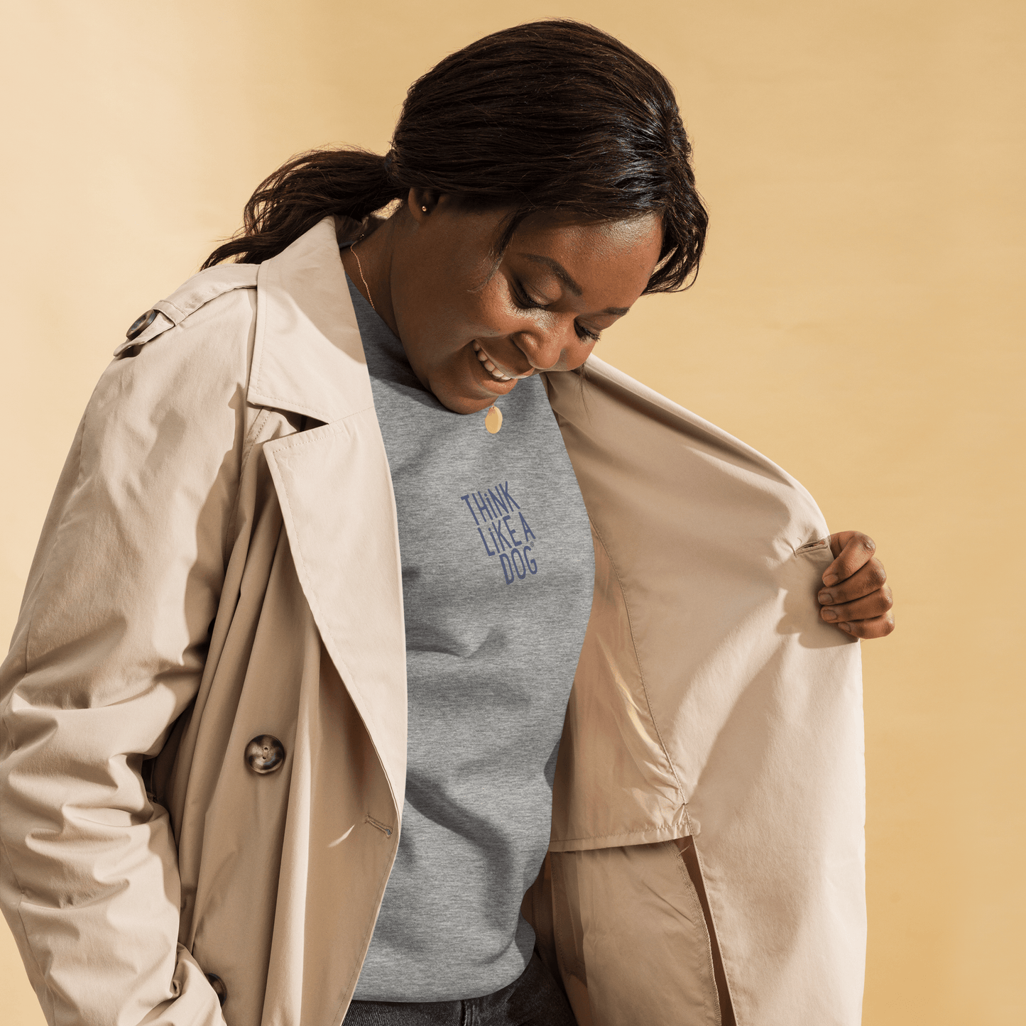 A woman in a beige trench coat smiles while showing off the THiNK LiKE A DOG® "Think Like a Dog Sweatshirt – Carbon Grey & Navy Logo Comfort," the ideal gift for any dog lover.