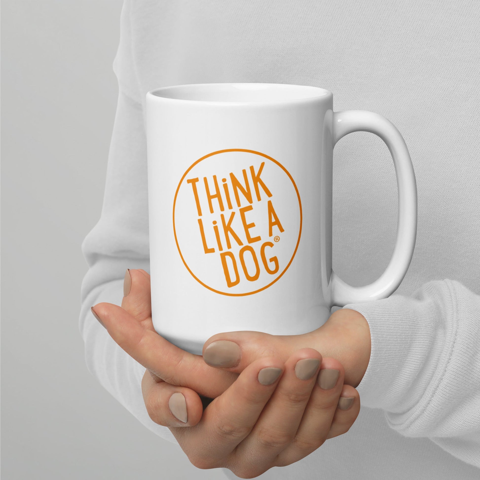 A person in a light sweater holds the THiNK LiKE A DOG® Think Like A Dog Mug – Vibrant Orange Logo, a white positivity coffee mug featuring "THiNK LiKE A DOG" in orange inside a circle.