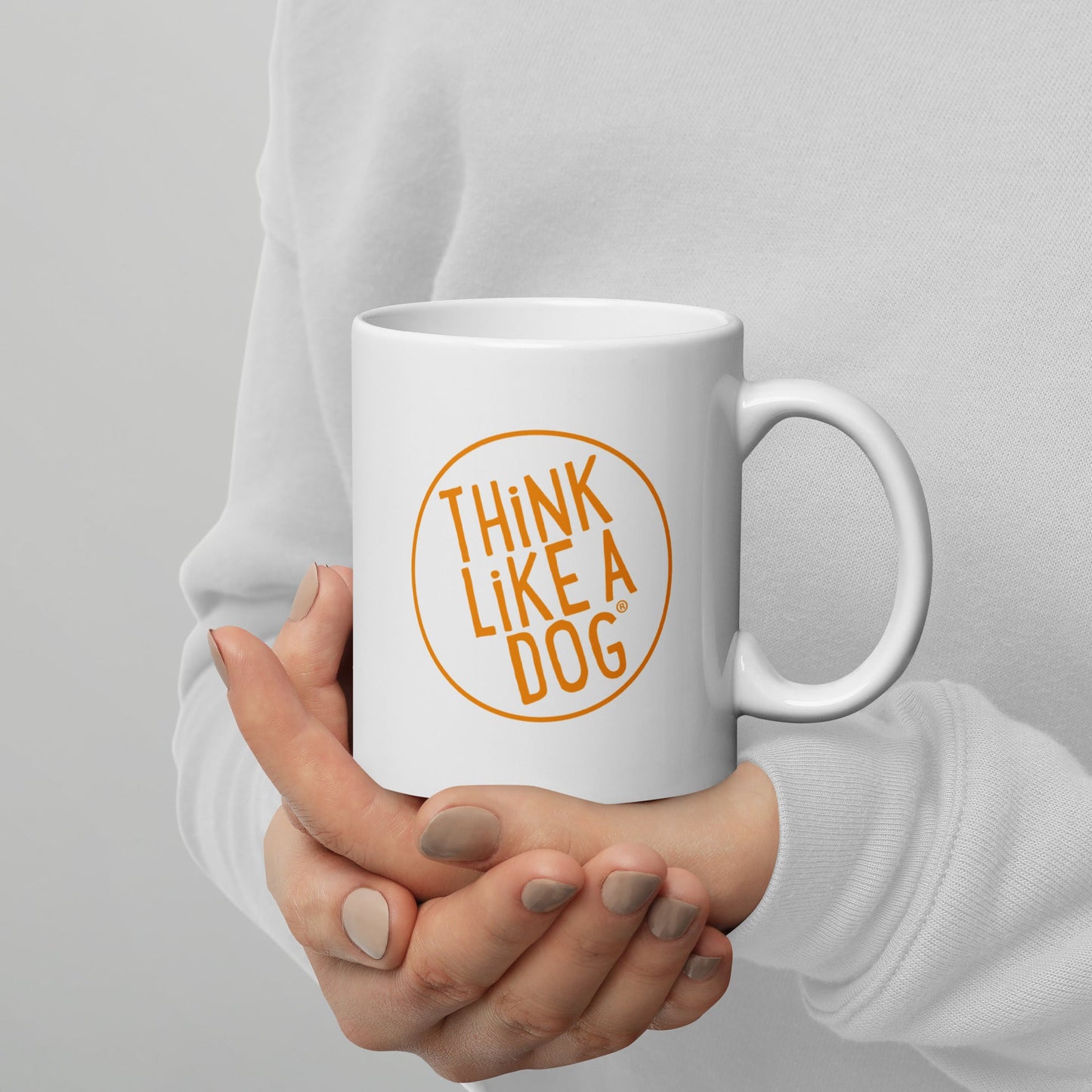 A person holds the THiNK LiKE A DOG® Think Like A Dog Mug – Vibrant Orange Logo, a white ceramic coffee mug with "THINK LIKE A DOG" in bold orange inside a circle—an ideal gift for dog lovers.