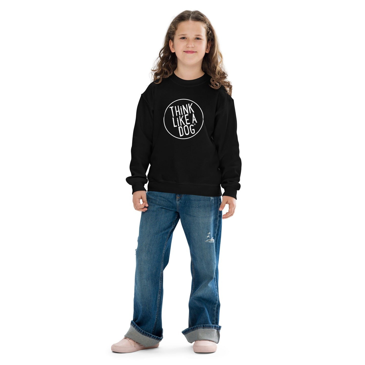 A young girl with shoulder-length curly hair is standing, wearing a THiNK LiKE A DOG® Kids' Cozy Crewneck Sweatshirt with White Think Like A Dog Logo – Cute and Comfortable Dog-Themed Apparel and blue jeans. She is looking towards the camera and smiling slightly.