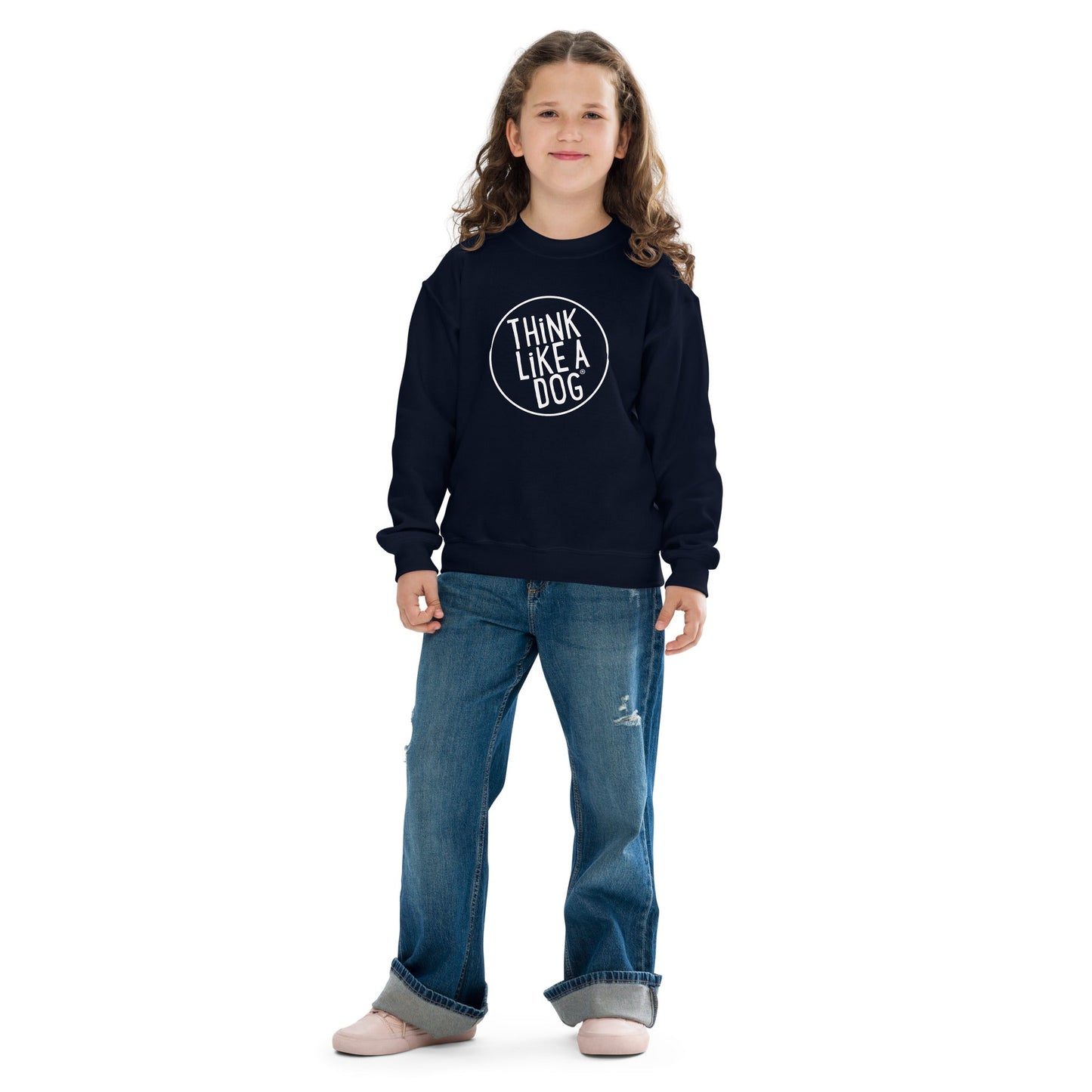 A young child with long curly hair stands wearing a high-quality Kids' Cozy Crewneck Sweatshirt with the white THiNK LiKE A DOG® logo in navy, paired with blue jeans, against a white background.