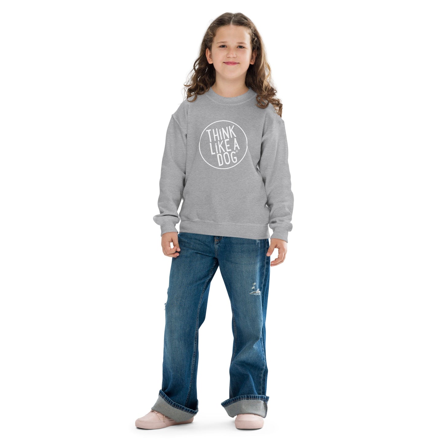 A young child with long hair wearing a Kids' Cozy Crewneck Sweatshirt with White Think Like A Dog Logo by THiNK LiKE A DOG®, paired with blue jeans, standing against a plain white background.