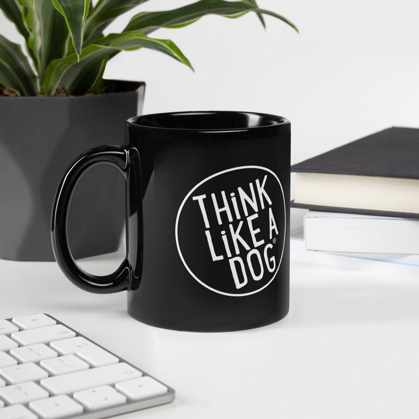 The THiNK LiKE A DOG® Black Mug – White Logo sits elegantly on a desk with a keyboard, books, and plant. Its minimalist design is perfect for any dog lover's workspace.