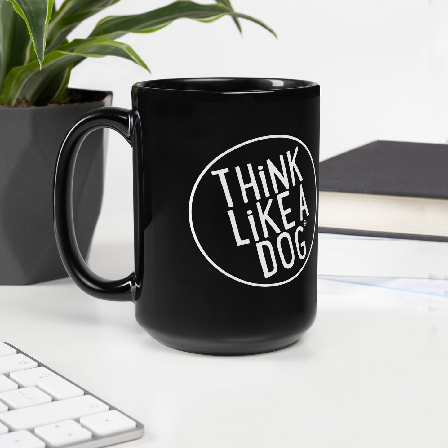The THiNK LiKE A DOG® Black Mug – White Logo, ideal for dog lovers, sits beside a plant, notebook, and keyboard on a white desk. Its minimalist design effortlessly enhances any workspace.