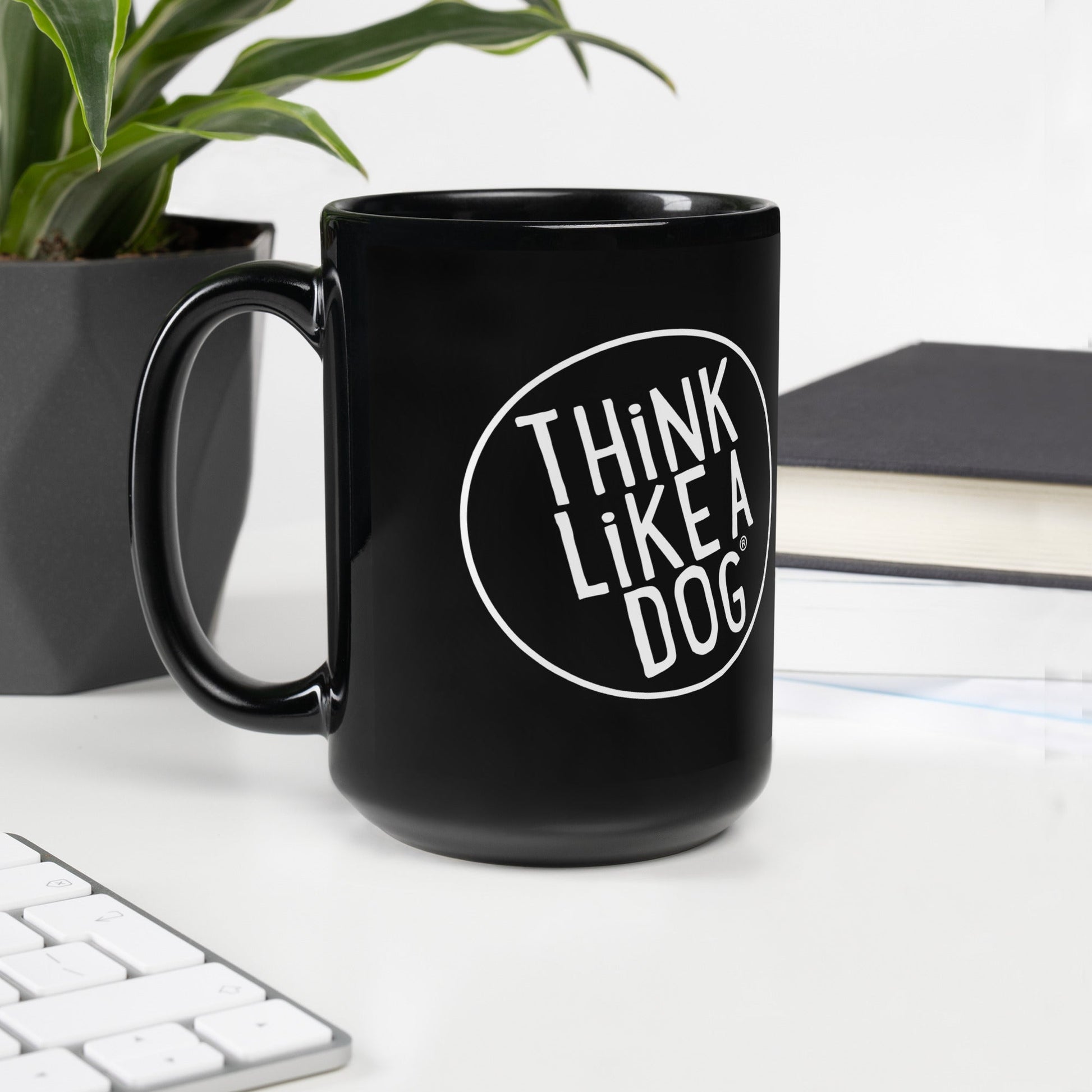 The THiNK LiKE A DOG® Black Mug – White Logo, ideal for dog lovers, sits beside a plant, notebook, and keyboard on a white desk. Its minimalist design effortlessly enhances any workspace.