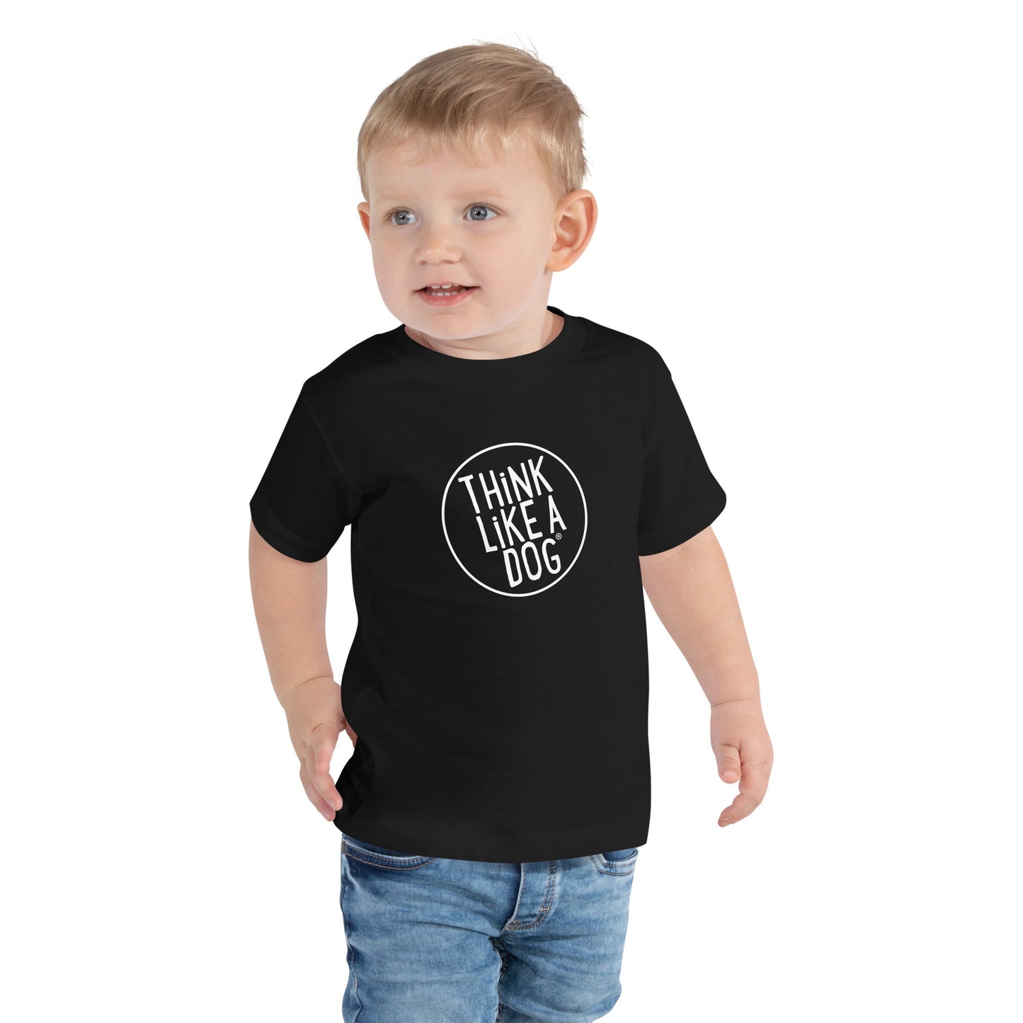 A joyful toddler in a THiNK LiKE A DOG® "THiNK LiKE A DOG" short sleeve tee, paired with blue jeans, poses against a plain backdrop. The ultra-soft and breathable ensemble from THiNK LiKE A DOG® ensures comfort while making a playful statement.