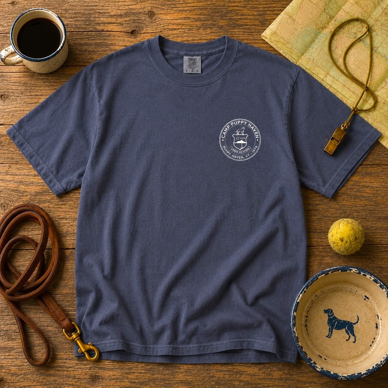 Camp Puppy Haven Badge Tee in Navy laid flat on dark wood with a leather leash, brass whistle, topo map, enamel mug of coffee, tennis ball, and a dog bowl