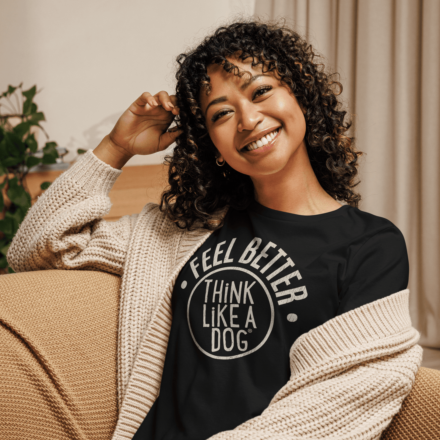 A curly-haired dog mom sits on a couch, wearing THiNK LiKE A DOG®'s ultra-soft "Feel Better Women's T-Shirt with Large Logo," perfectly paired with a cozy beige sweater.