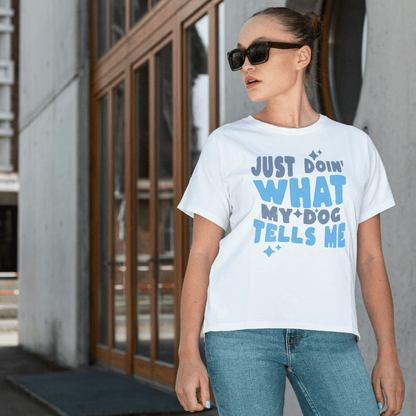 A woman in sunglasses and a white t-shirt from THiNK LiKE A DOG® with the text "Just Doin' What My Dog Tells Me" stands outside near wooden-framed windows, highlighting a humorous and charming message for dog lover shirt enthusiasts.