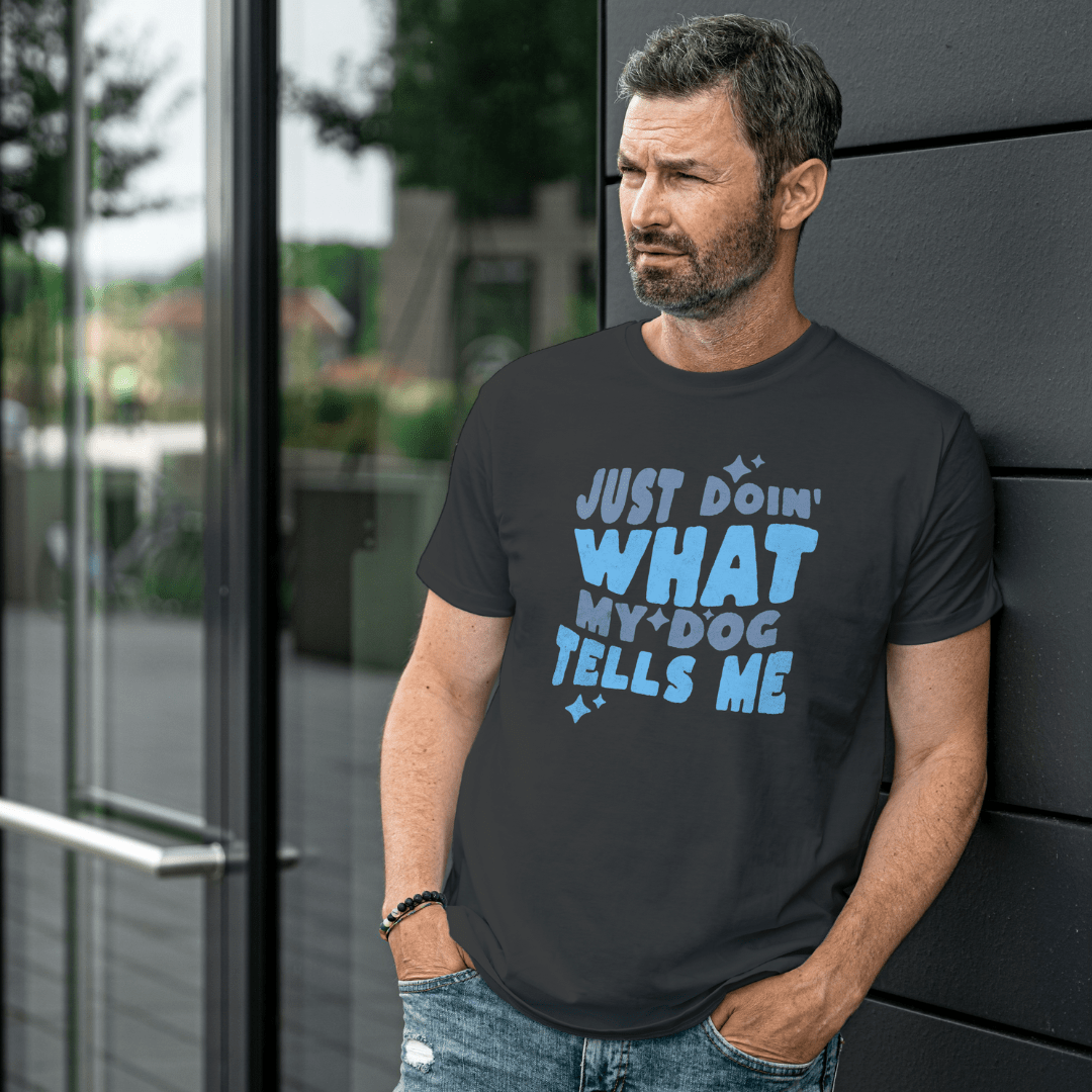 A man in a casual outfit leans against a wall, wearing the "Just Doin' What My Dog Tells Me" Funny Dog Lover Shirt by THiNK LiKE A DOG®. It's the perfect humorous shirt for any dog lover.