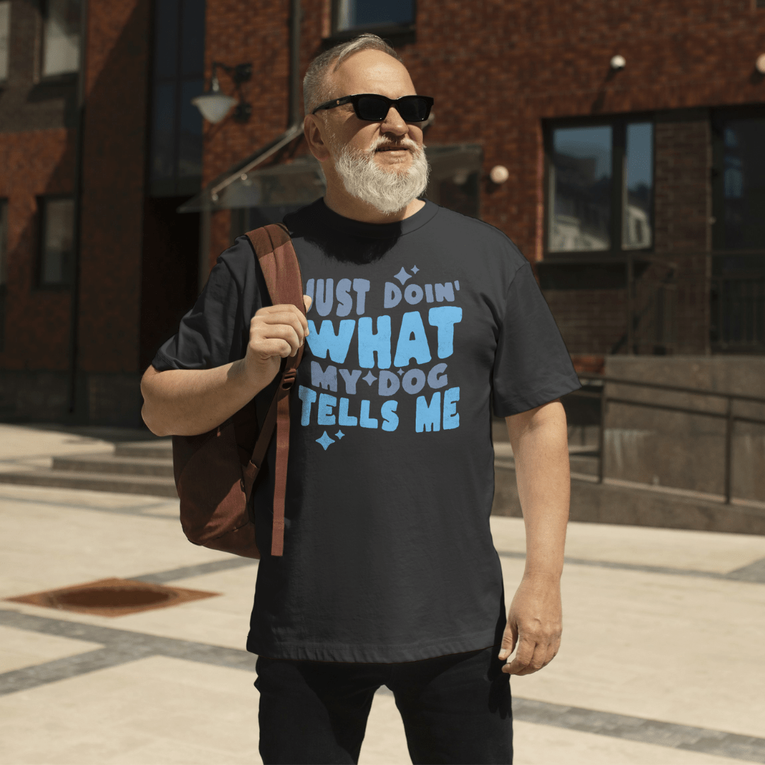 A man in sunglasses and casual attire wears the THiNK LiKE A DOG® Funny Dog Lover Shirt saying, "Just Doin' What My Dog Tells Me," while standing outside a brick building, holding a backpack.