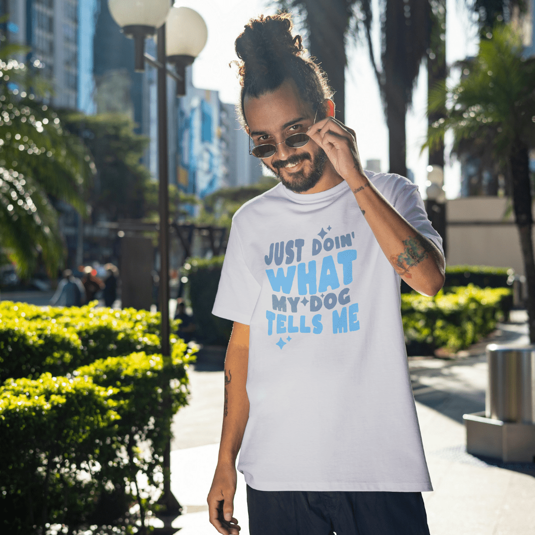 A man in sunglasses and a white THiNK LiKE A DOG® T-shirt with the funny dog-themed message "Just Doin' What My Dog Tells Me" stands on a sunny, tree-lined street. Ideal for anyone wanting humorous shirts that showcase their love for dogs in style.