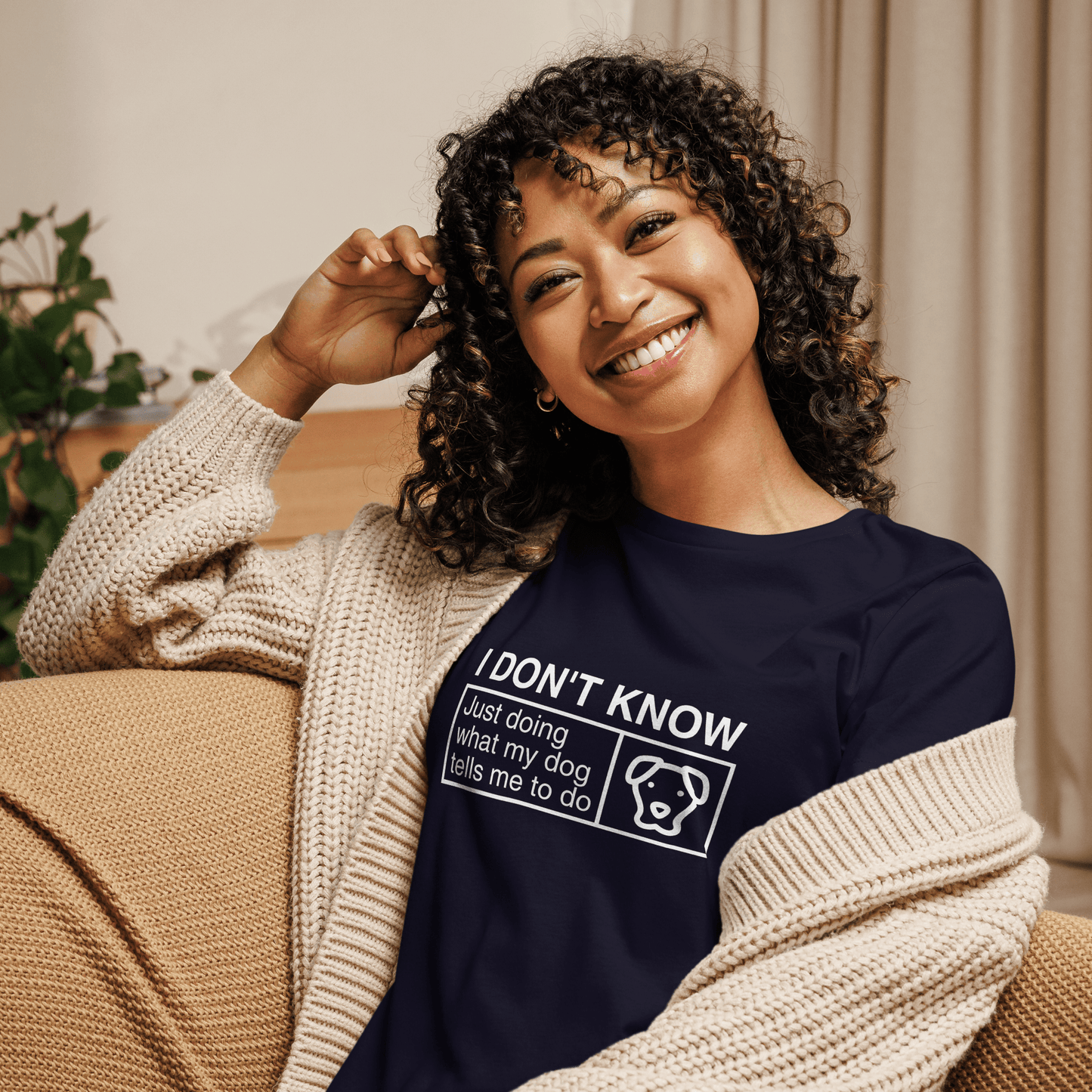 A smiling person with curly hair sits on a couch, wearing an open cardigan and THiNK LiKE A DOG®'s navy Funny Dog Shirt that says, "Just Doing What My Dog Tells Me.