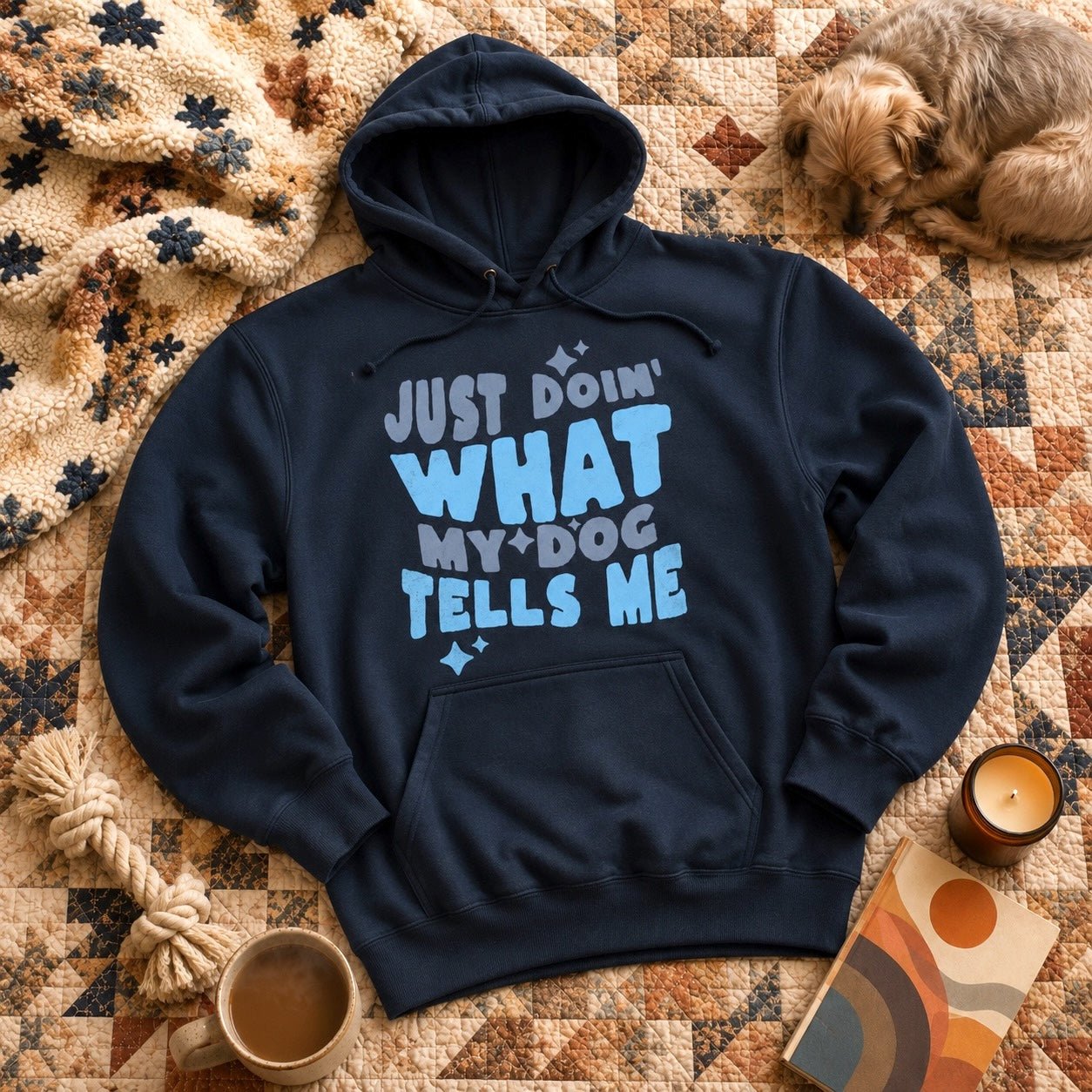 Navy "Just Doin' What My Dog Tells Me" hoodie laid flat on a quilted blanket with a sleeping terrier, sherpa throw, candle, coffee mug, and rope toy
