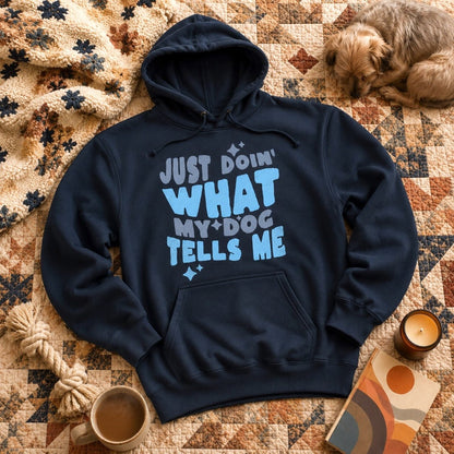 Navy "Just Doin' What My Dog Tells Me" hoodie laid flat on a quilted blanket with a sleeping terrier, sherpa throw, candle, coffee mug, and rope toy