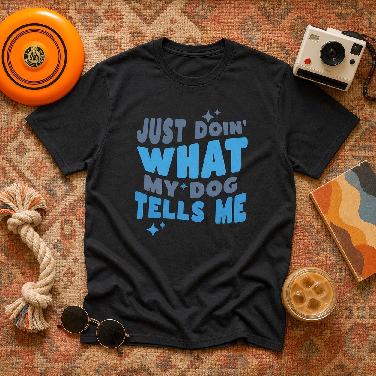 Black "Just Doin' What My Dog Tells Me" t-shirt in a flat lay with an orange frisbee, Polaroid camera, rope dog toy, round sunglasses, and iced coffee on a vintage patterned rug